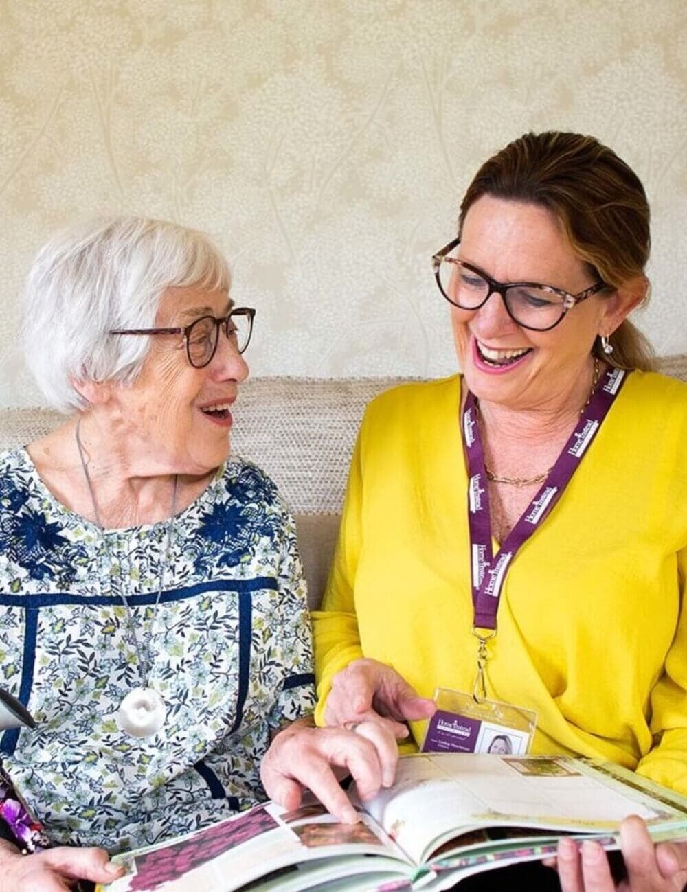 An elderly woman and a younger woman with glasses, both smiling and looking at a book together. - Home Instead