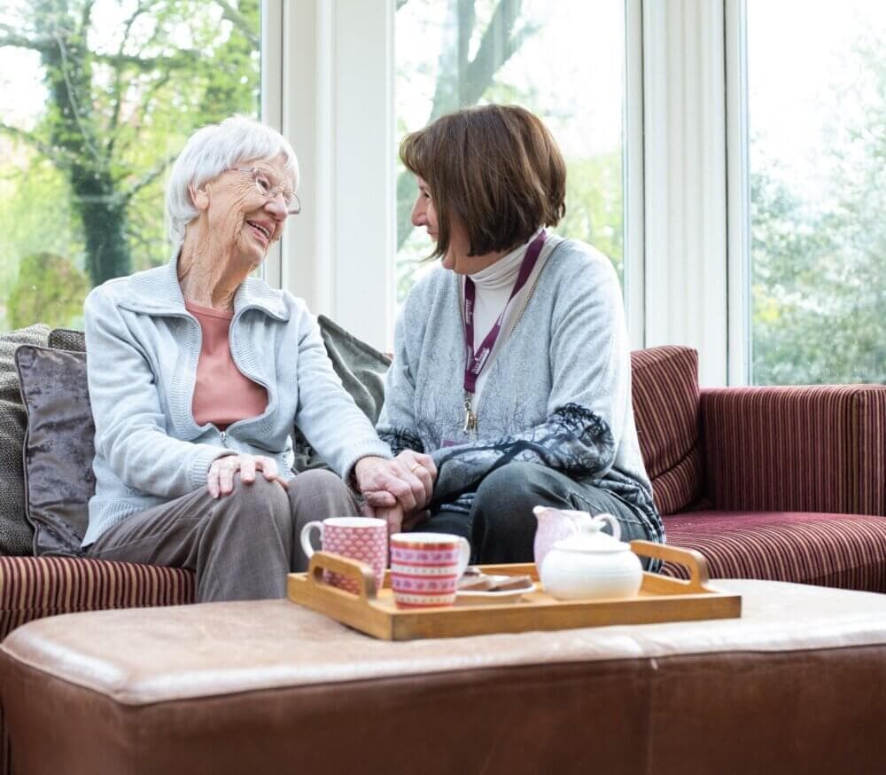 An elderly woman and a younger woman sit together, holding hands and smiling, with tea on a tray in front of them. - Home Instead