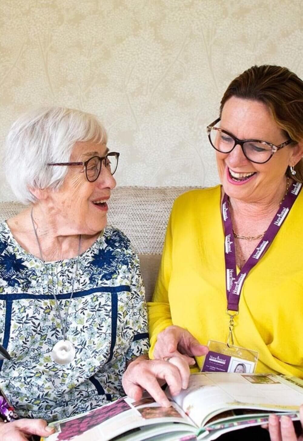 Elderly woman and younger woman in yellow shirt, both wearing glasses, smiling and looking at a book together. - Home Instead