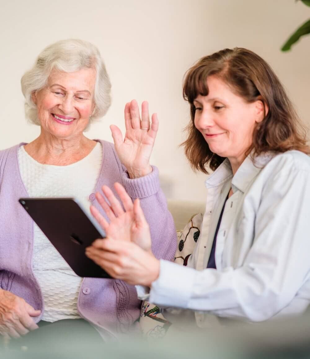 Older woman and younger woman smiling and waving at a tablet screen during a video call. - Home Instead