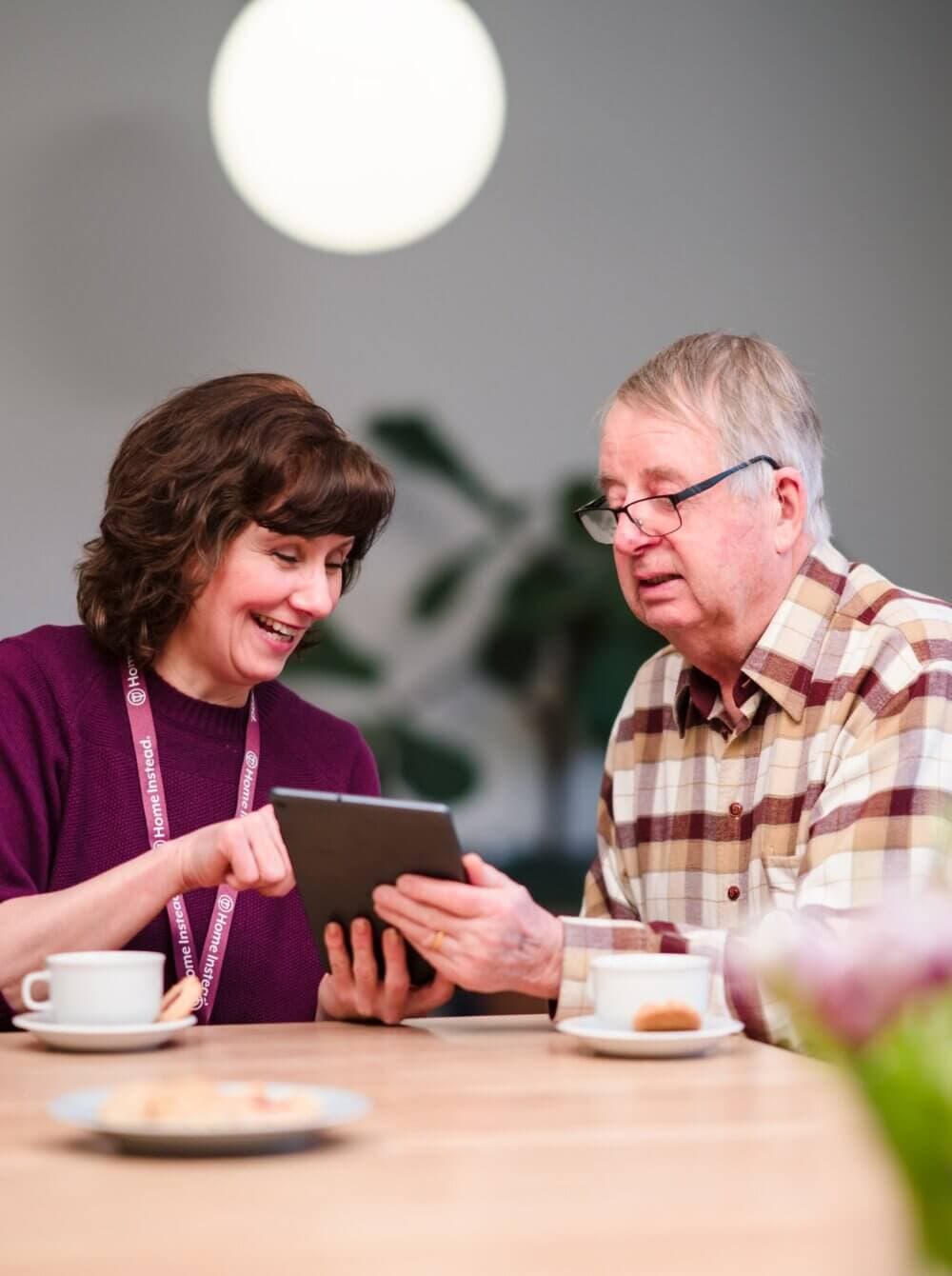 A woman shows an older man something on a tablet at a table with teacups and flowers. - Home Instead