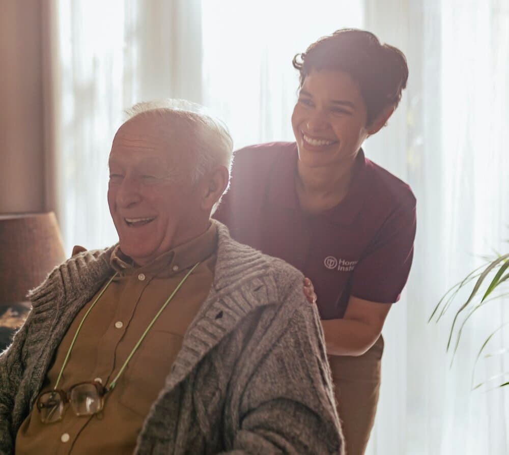 A caregiver cheerfully tends to a smiling elderly man sitting in a chair, with sunlight streaming in behind them. - Home Instead