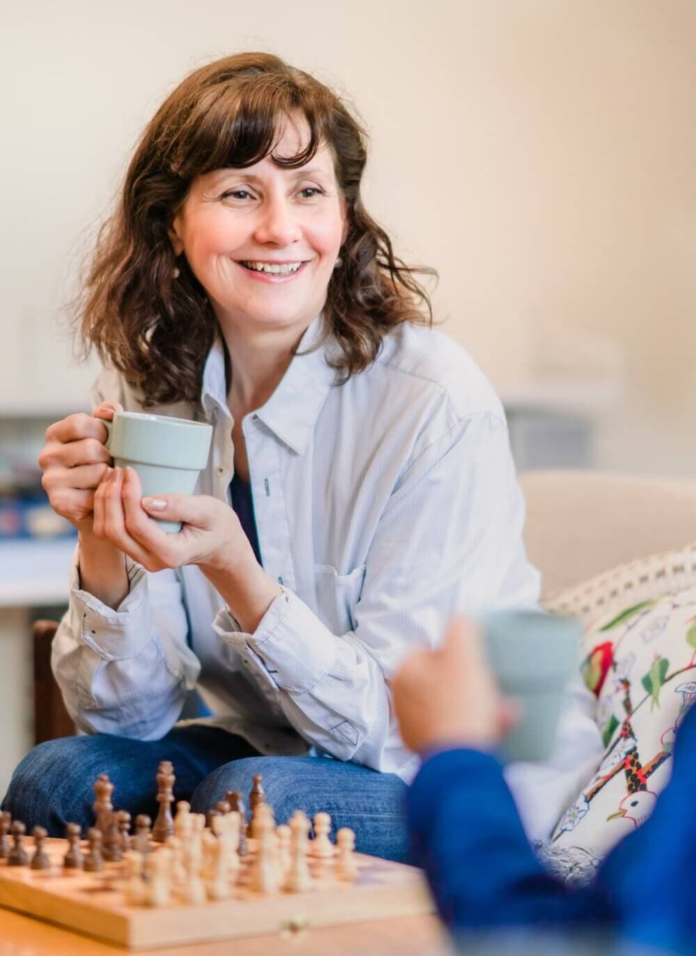 Smiling woman holding a mug, sitting opposite someone at a table with a chessboard. - Home Instead