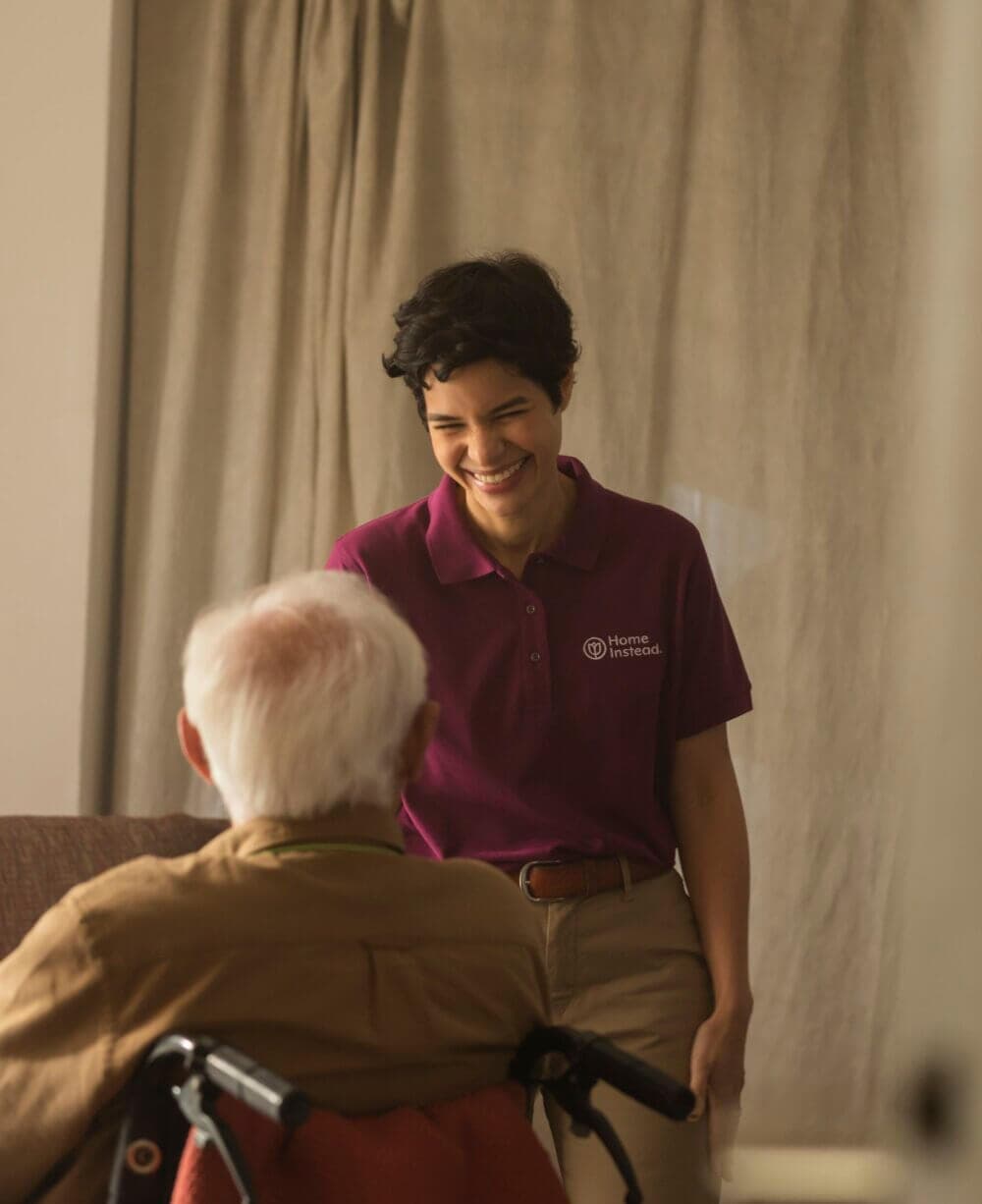 Caregiver in a purple shirt smiling and talking with an elderly person in a wheelchair, indoors. - Home Instead