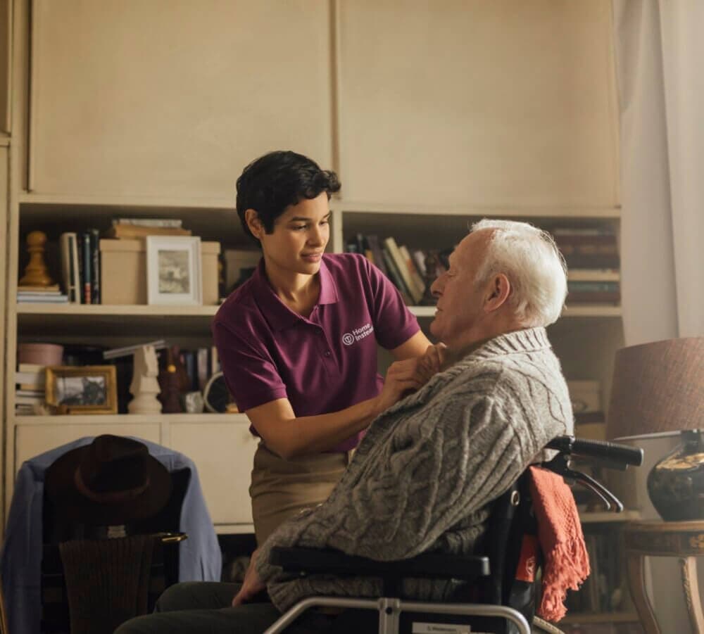 A caregiver in a purple shirt assists an elderly man sitting in a wheelchair within a warmly lit room. - Home Instead