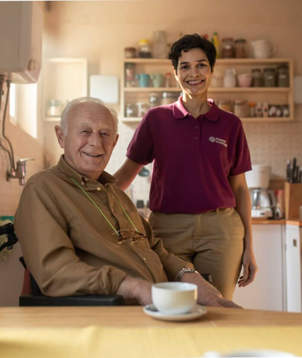 An elderly man in a wheelchair sits beside a smiling caregiver in a kitchen, with shelves of dishes in the background. - Home Instead