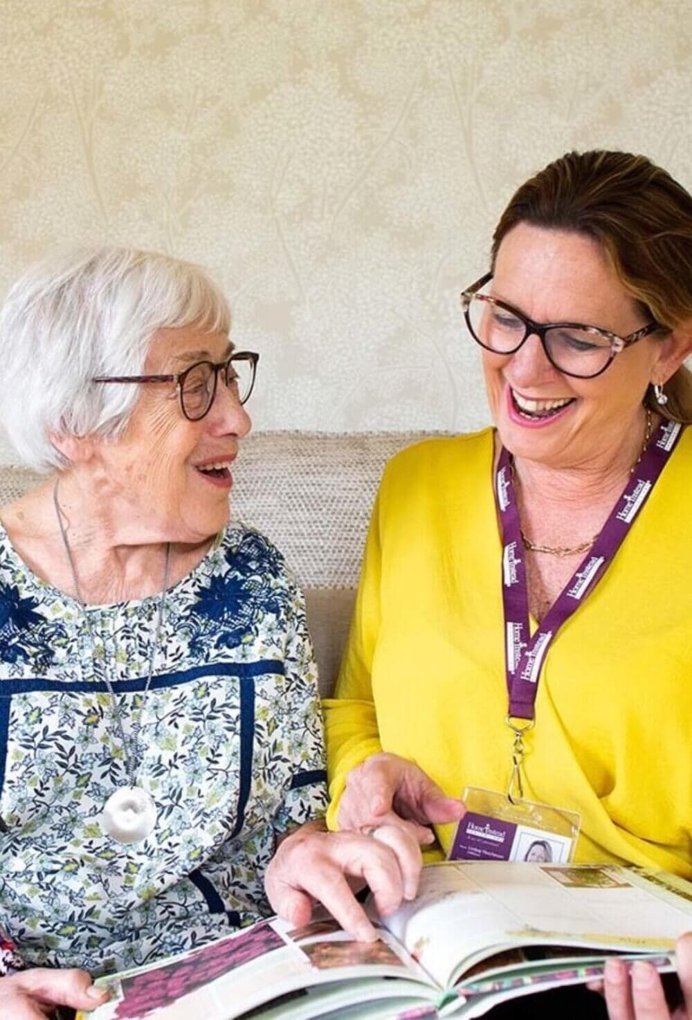 An elderly woman and a caregiver with glasses and a lanyard are smiling while looking at a book together. - Home Instead