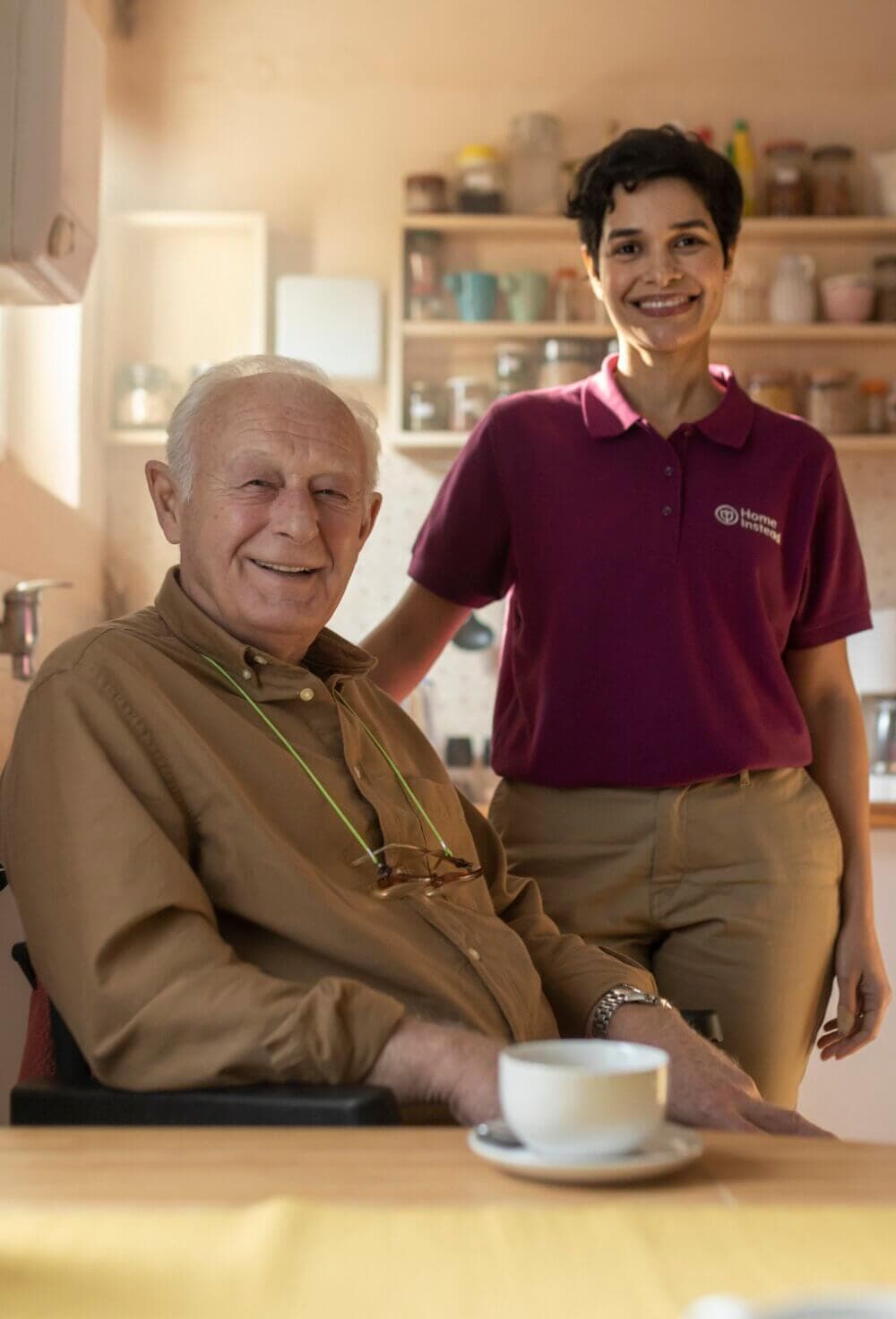 An elderly man seated at a table with a cup of coffee, and a woman in a purple shirt standing behind him, both smiling. - Home Instead