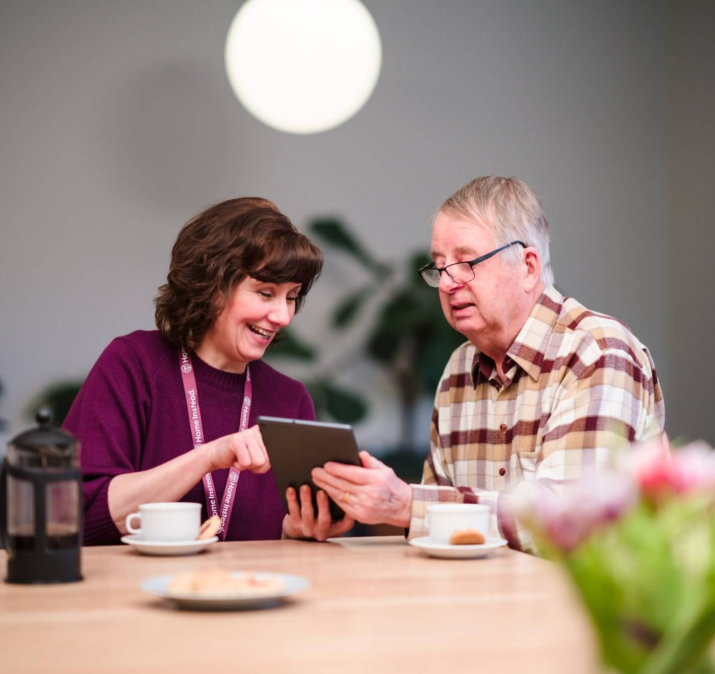 A woman and an elderly man share a tablet screen, smiling, with coffee and pastries on the table in front of them. - Home Instead