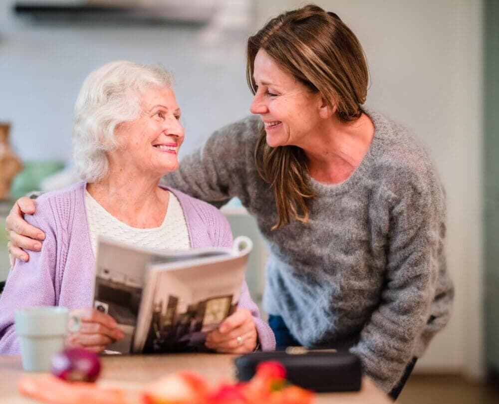 Smiling older woman holding a magazine, and a younger woman embracing her from the side in a cozy home setting. - Home Instead