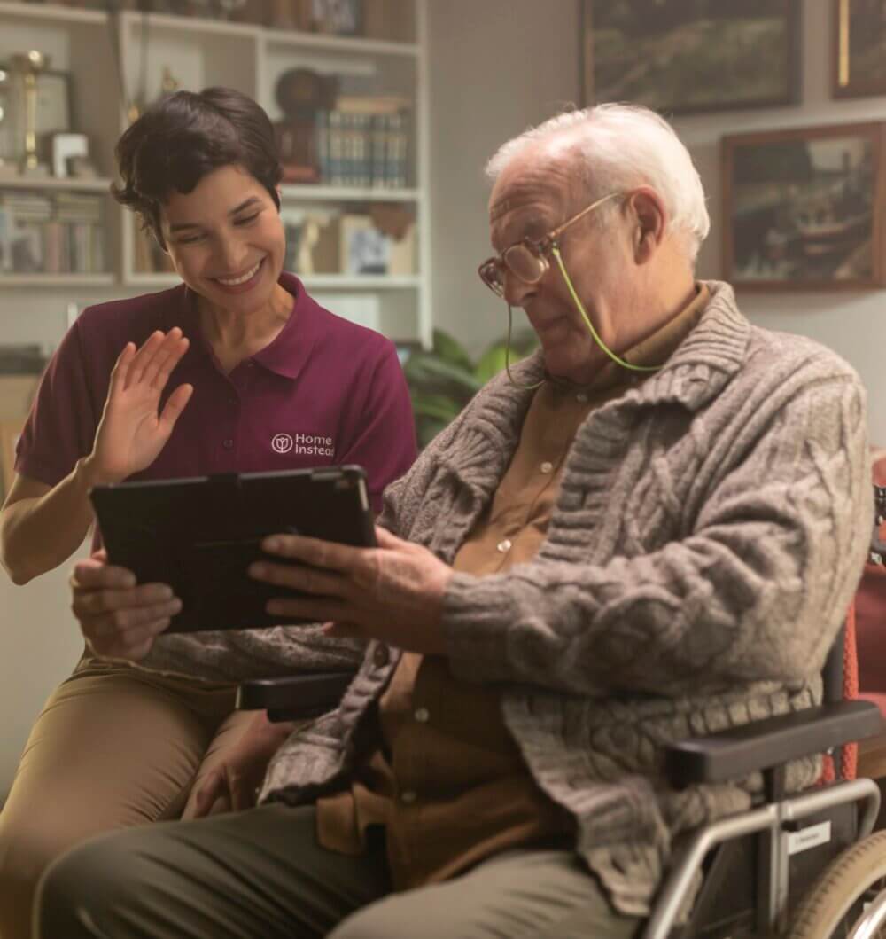 A caregiver smiles and waves while assisting an elderly man in a wheelchair who is looking at a tablet. - Home Instead