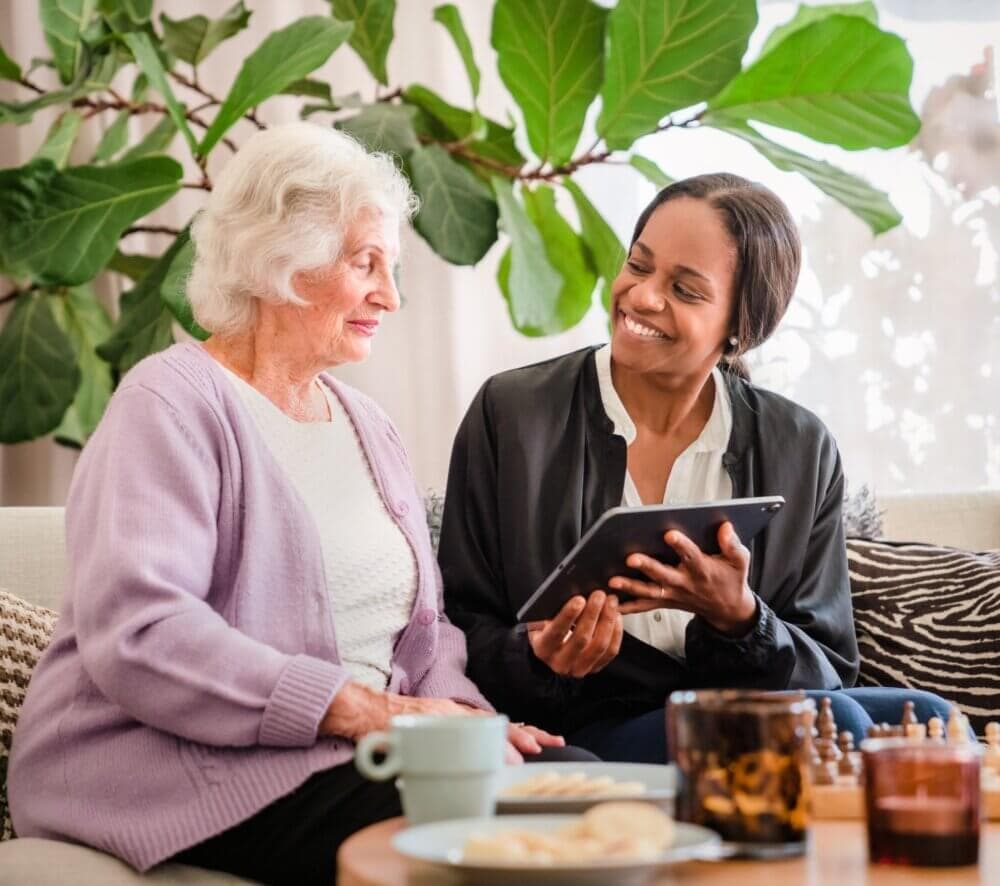 A young woman shows something on a tablet to an older woman sitting next to her, both smiling, with a plant in the background. - Home Instead