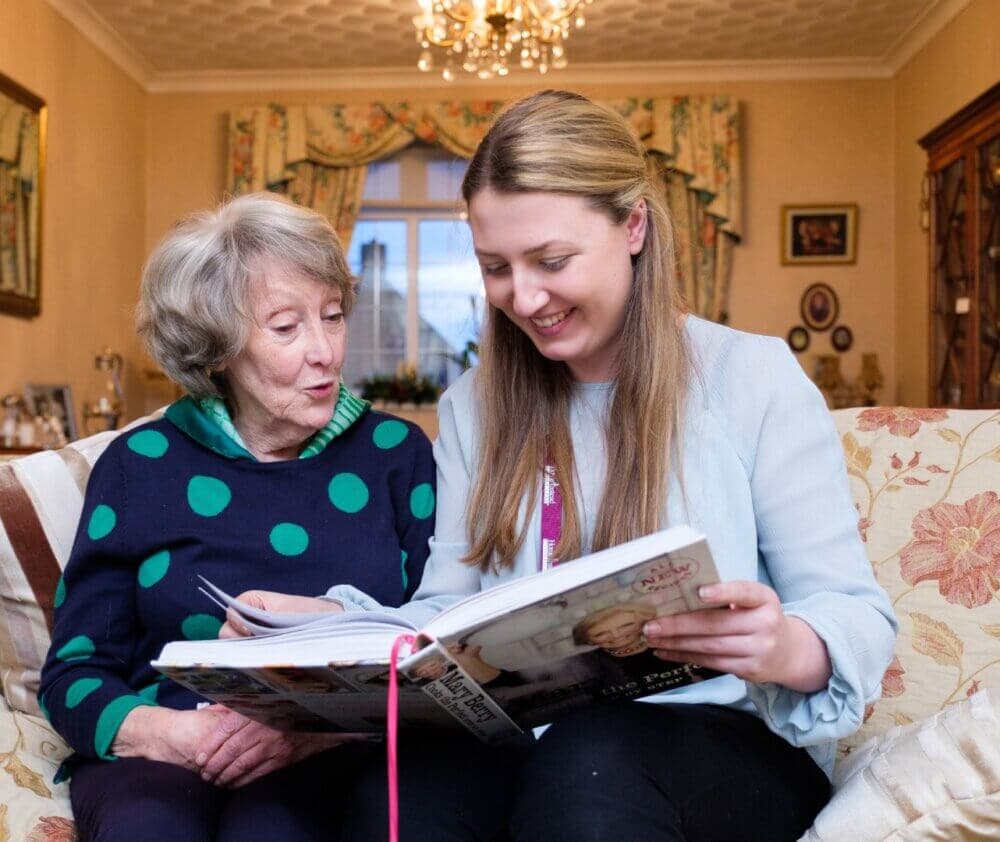 An elderly woman and a young woman smiling as they look through a book together in a cozy living room. - Home Instead