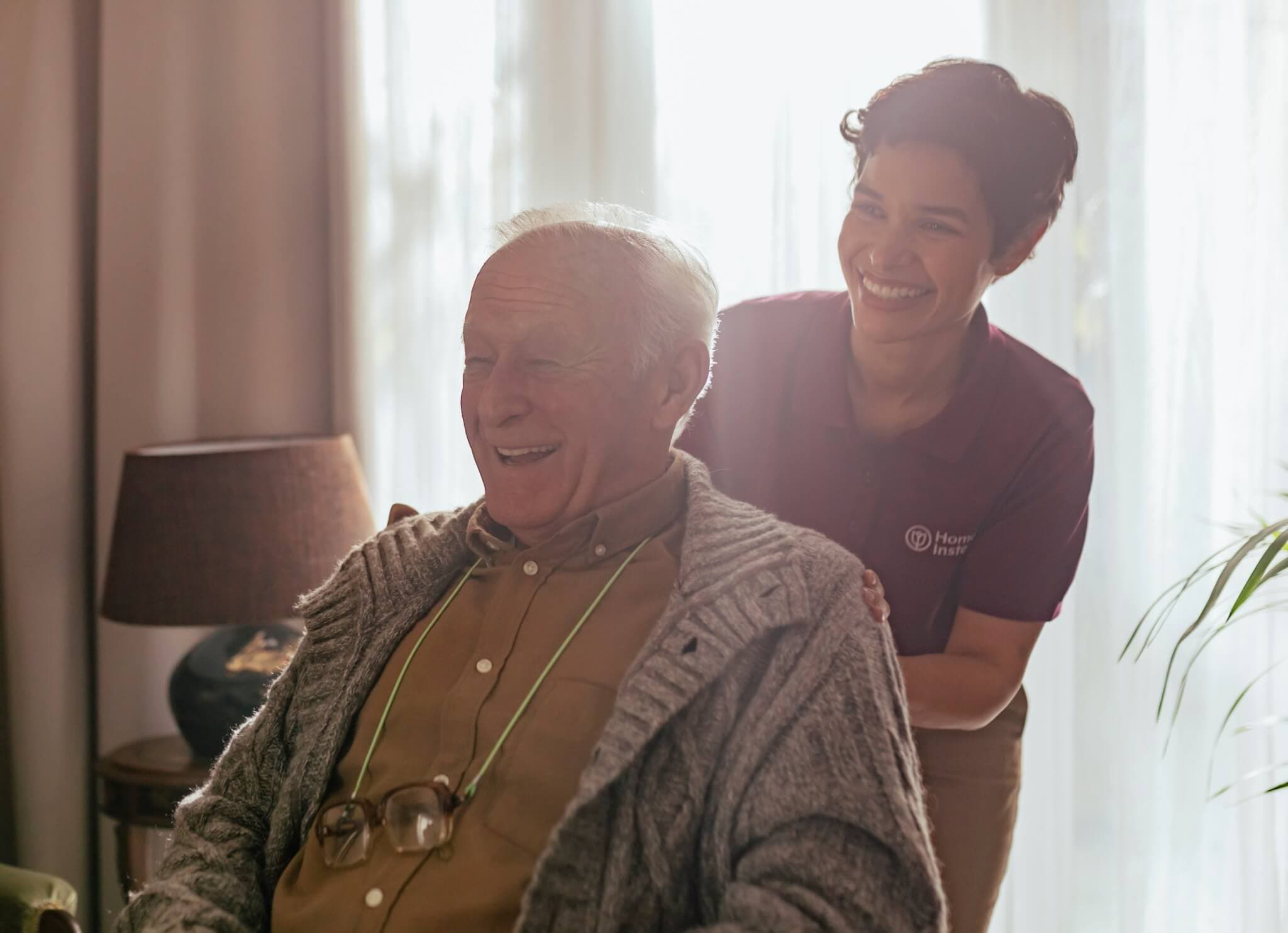 Home Instead Bournemouth and Christchurch Care Professional smiling and assisting an elderly man in a cosy room with sunlight in the background.