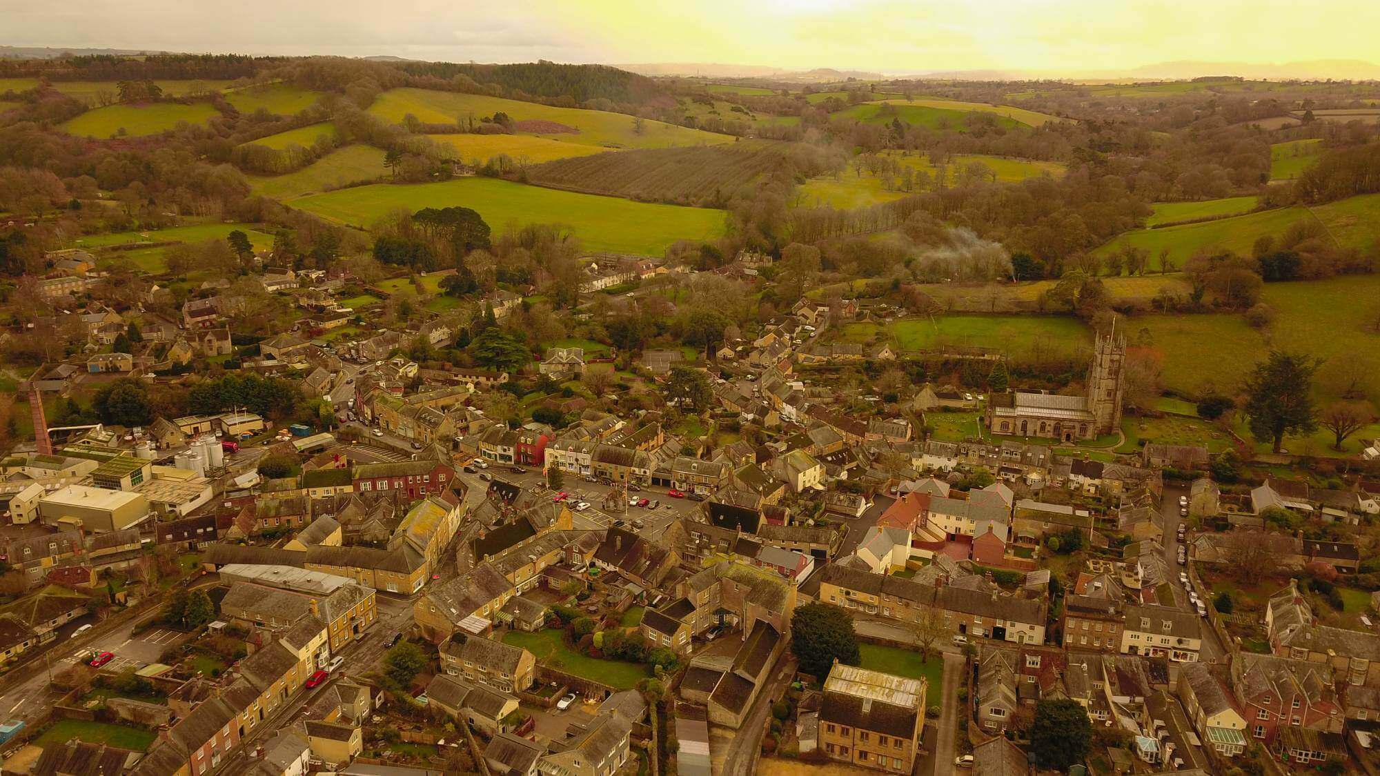 Aerial view of a small town with winding roads, buildings, and surrounding green hills during sunset. - Home Instead