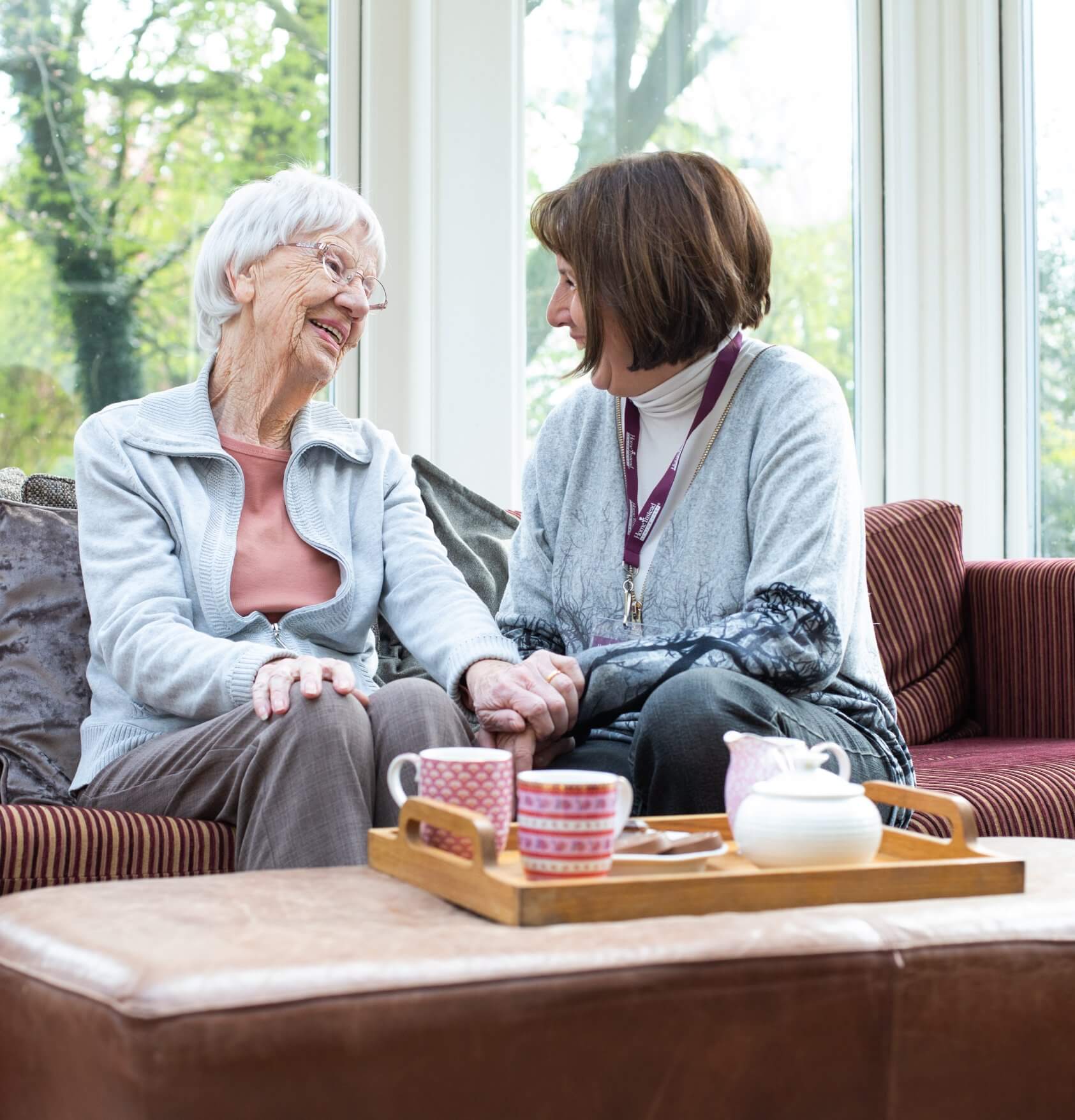 An elderly woman and a caregiver share a smile and hold hands while sitting on a cozy couch with tea on a tray. - Home Instead
