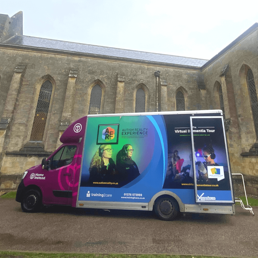 A parked van decorated with "Autism Reality Experience" and "Virtual Dementia Tour" graphics, in front of a historic building. - Home Instead