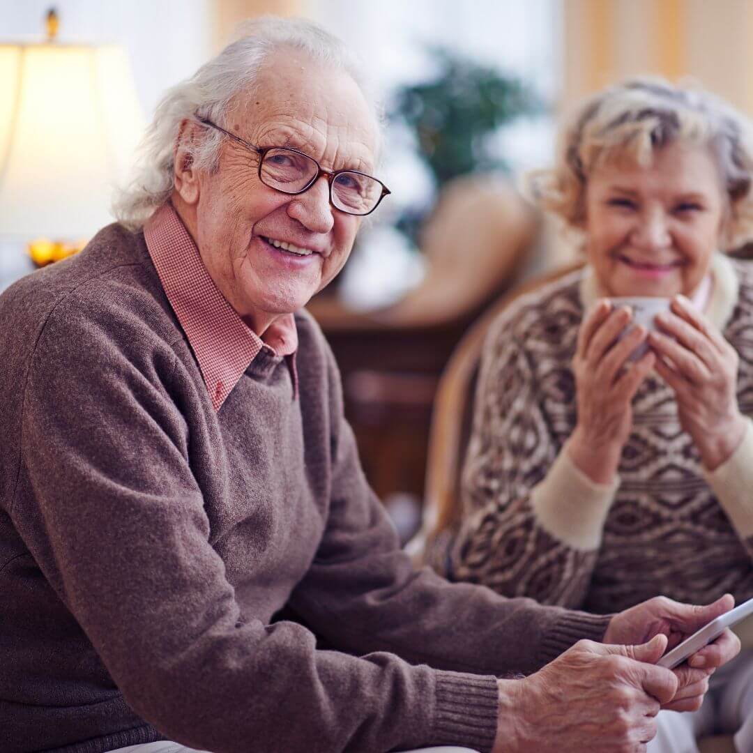 Elderly man with glasses smiling and holding a tablet, sitting next to a woman with a cup, both in cozy sweaters. - Home Instead