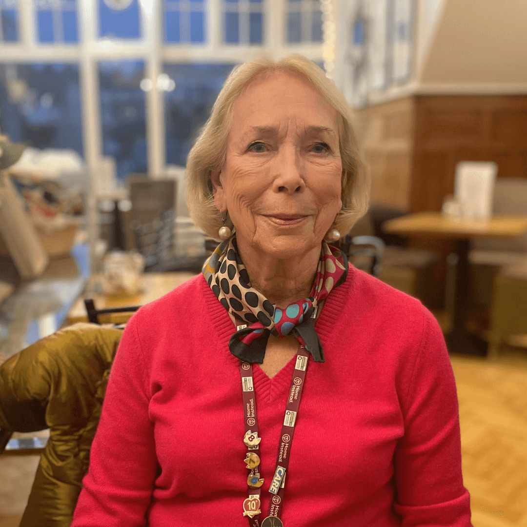 An elderly woman in a pink sweater and polka dot scarf smiles while seated in a cozy room with large windows. - Home Instead
