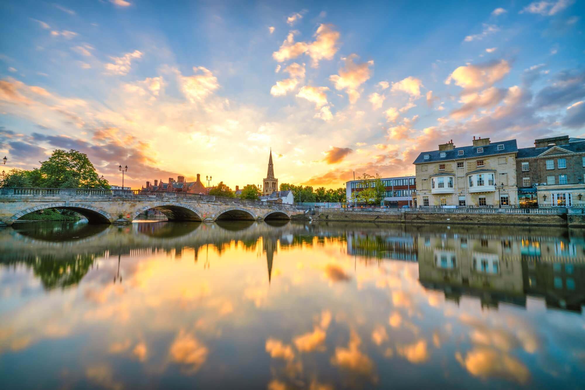 Sunset over a serene river with a historic bridge, church spire, and charming buildings reflecting in the water. - Home Instead
