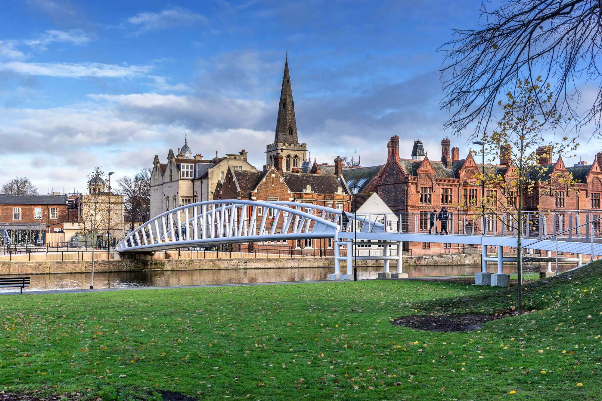 A white pedestrian bridge over a river, with historic buildings and a church spire in the background on a partly cloudy day. - Home Instead