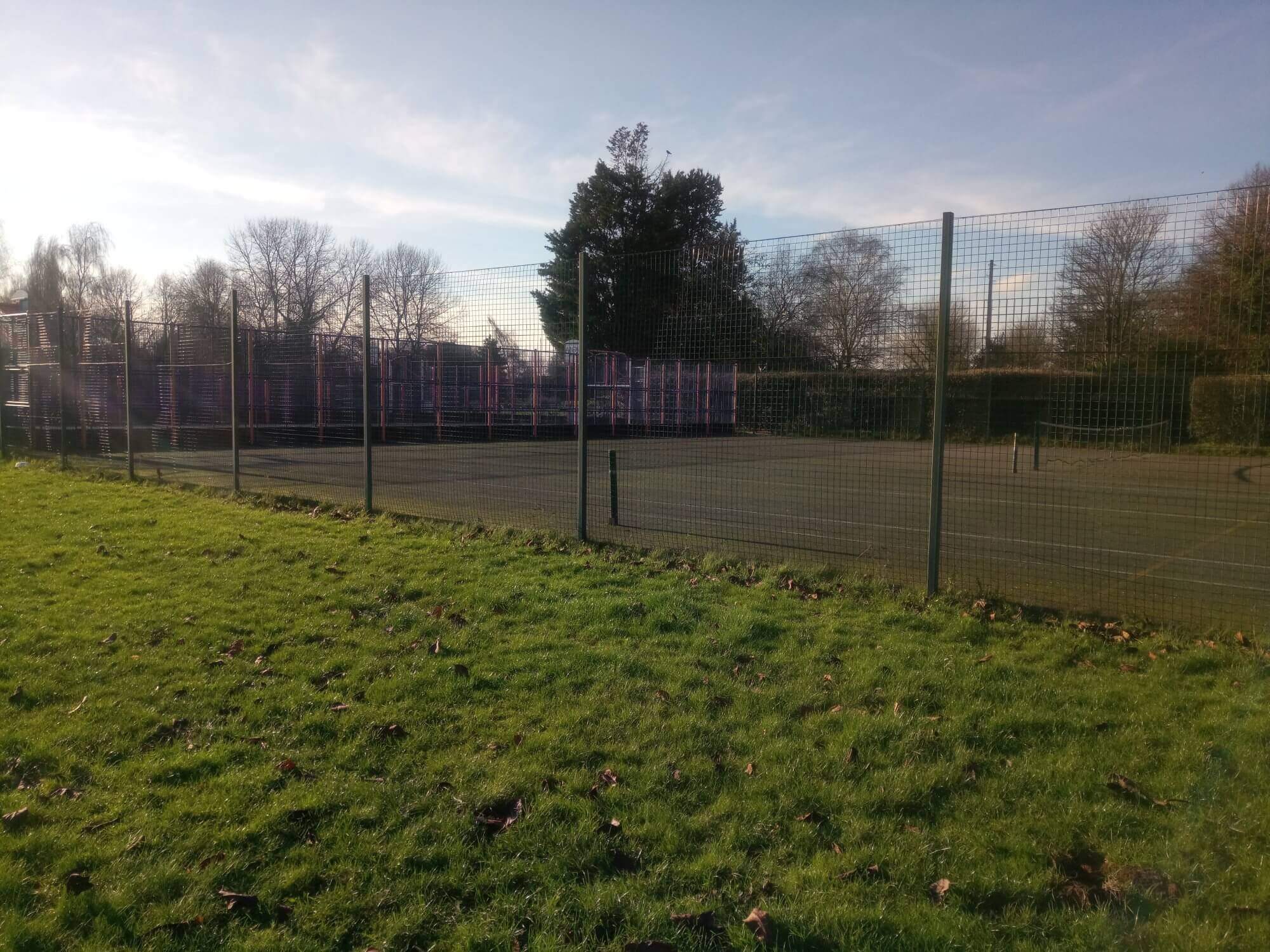 A fenced outdoor tennis court with trees and grass surrounding it on a sunny day. - Home Instead