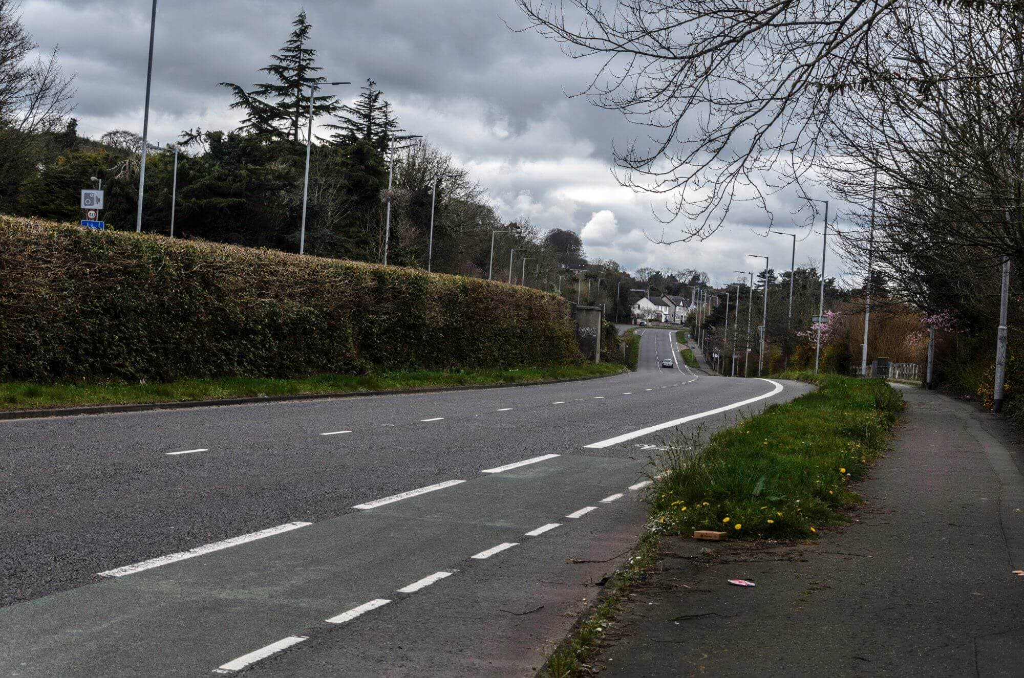 A deserted bending road lined with trees and hedges under a cloudy sky, with a sidewalk and bike lane on the right. - Home Instead