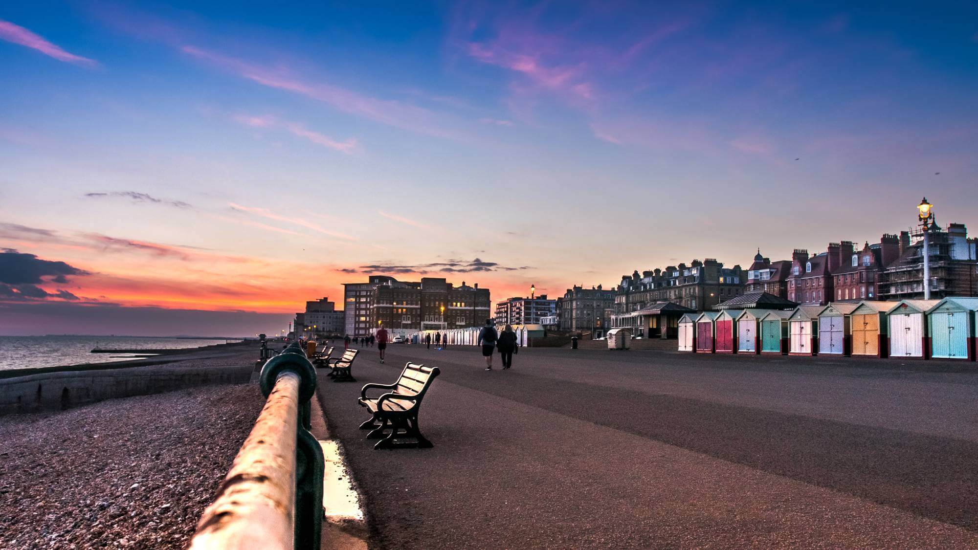 Sunset over a promenade with benches, beach huts, and people walking along the coast. Historic buildings in the background. - Home Instead