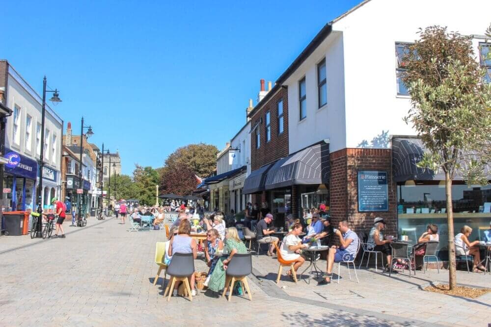 People dining and walking on a bustling, sunny street with shops and outdoor seating in front of cafes. - Home Instead