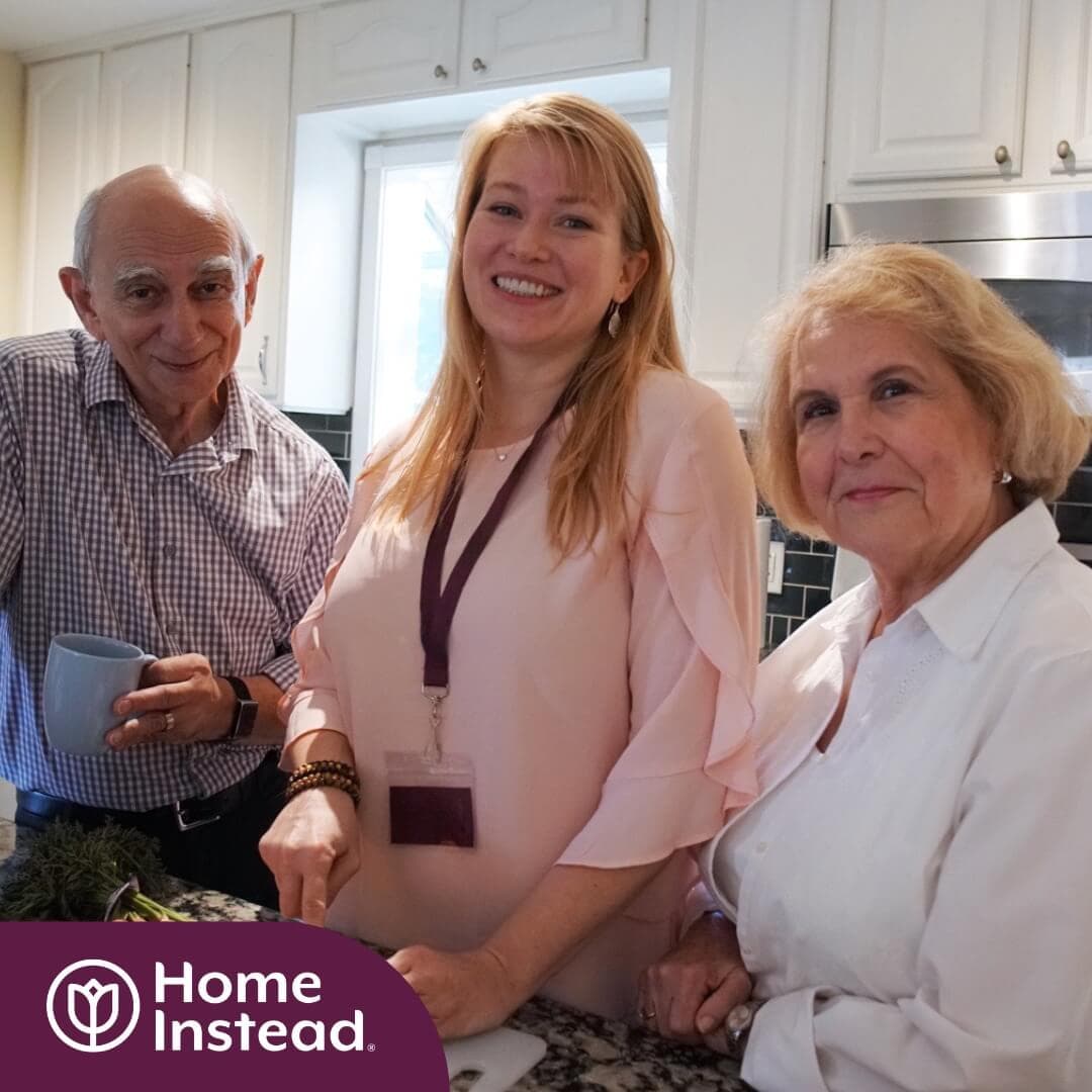 Three people smile in a kitchen with a "Home Instead" logo in the corner. One woman wears a lanyard. - Home Instead