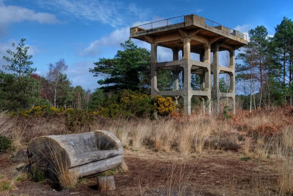 Abandoned concrete structure with a wooden log bench in a grassy, wooded area under a blue cloudy sky. - Home Instead