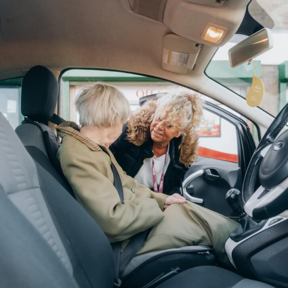 An elderly woman sits in a car while a caregiver leans in from outside, talking to her with care and attention. - Home Instead
