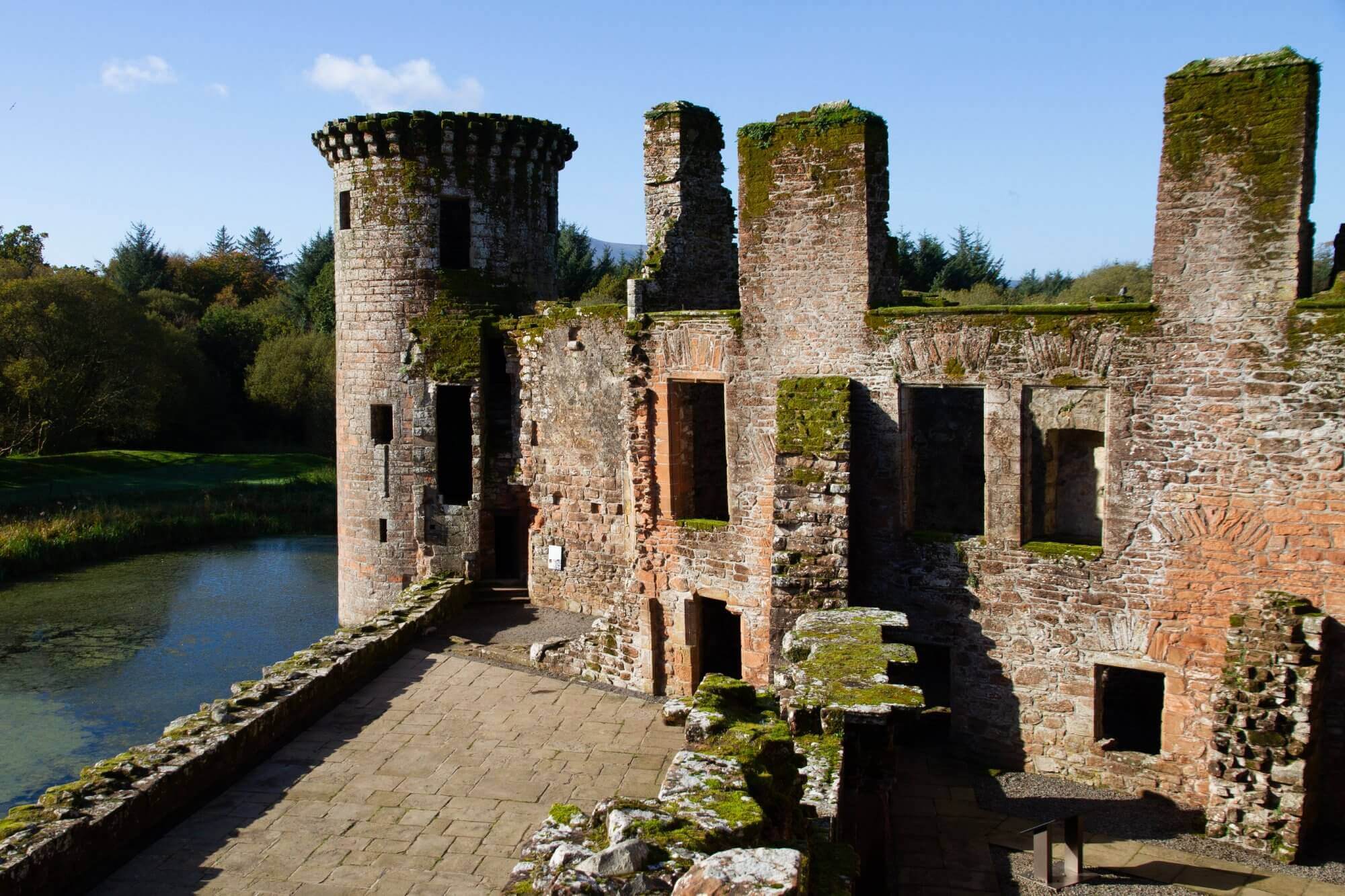 Ruins of an old stone castle with a round tower, surrounded by greenery and a small river. - Home Instead