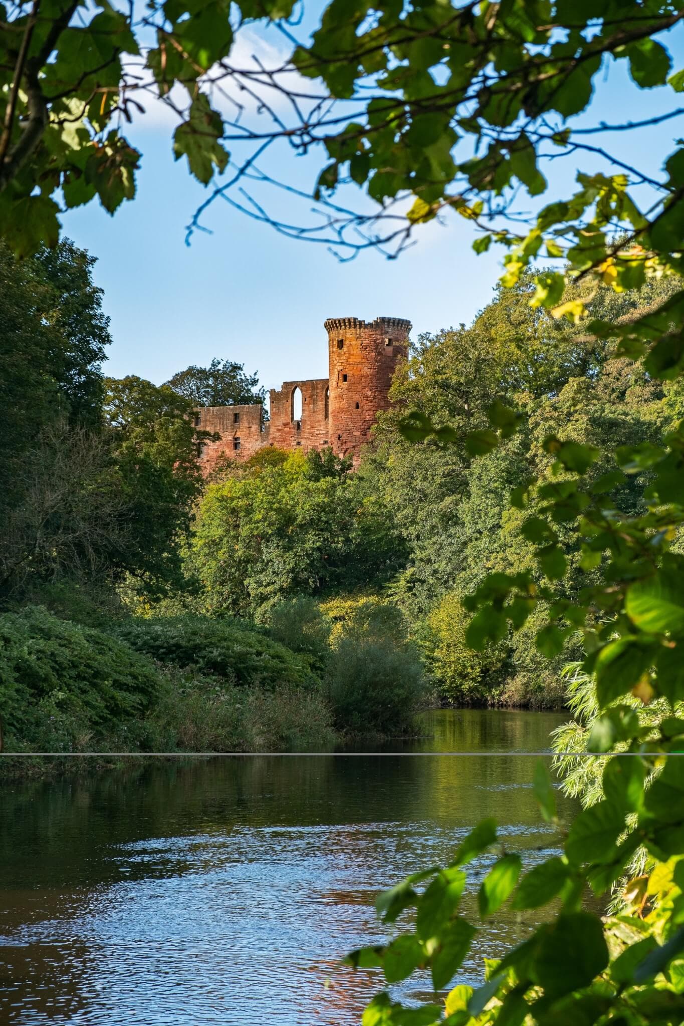 Red-brick castle towers peek through lush green foliage, overlooking a serene river under a clear sky. - Home Instead