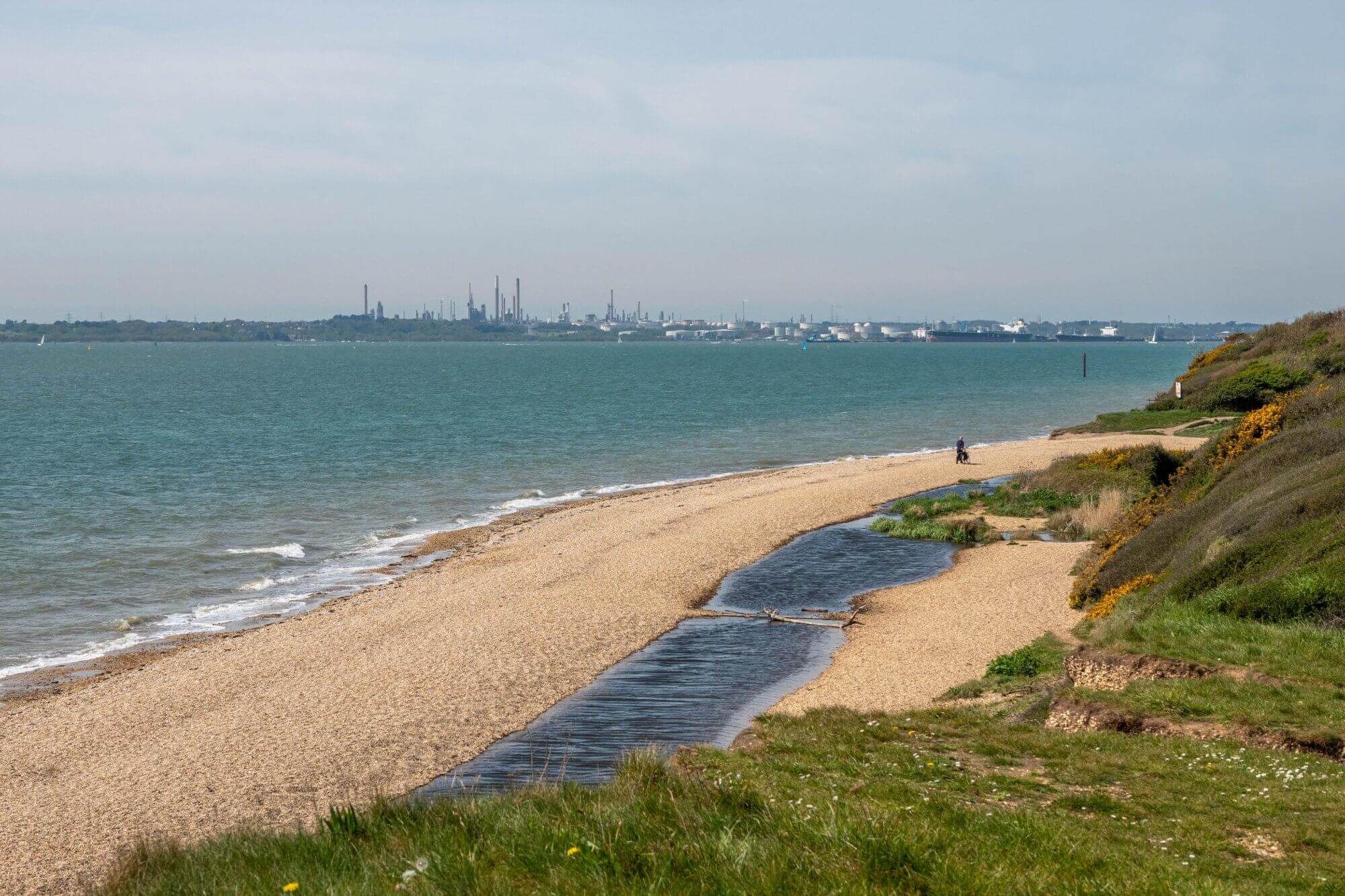 A serene beach with calm waves, a figure walking along the shore, and industrial buildings visible in the distance. - Home Instead