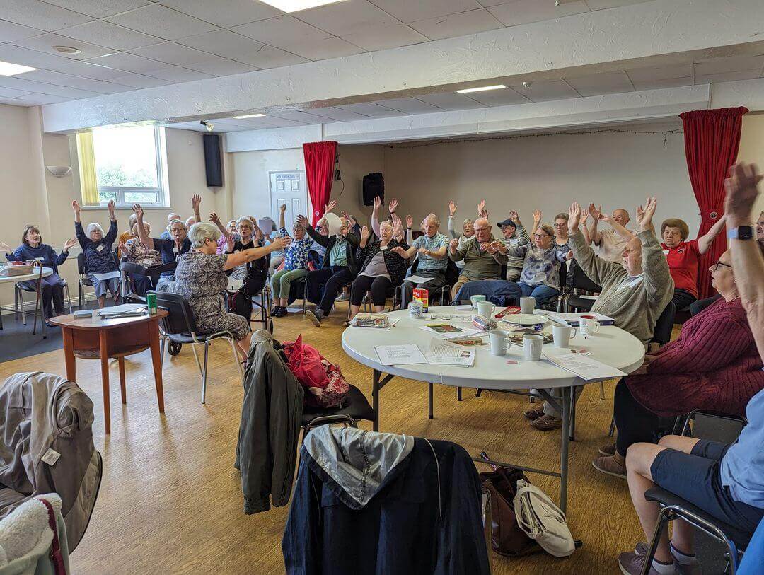 A group of elderly people sitting in a room with hands raised, participating in an activity. - Home Instead