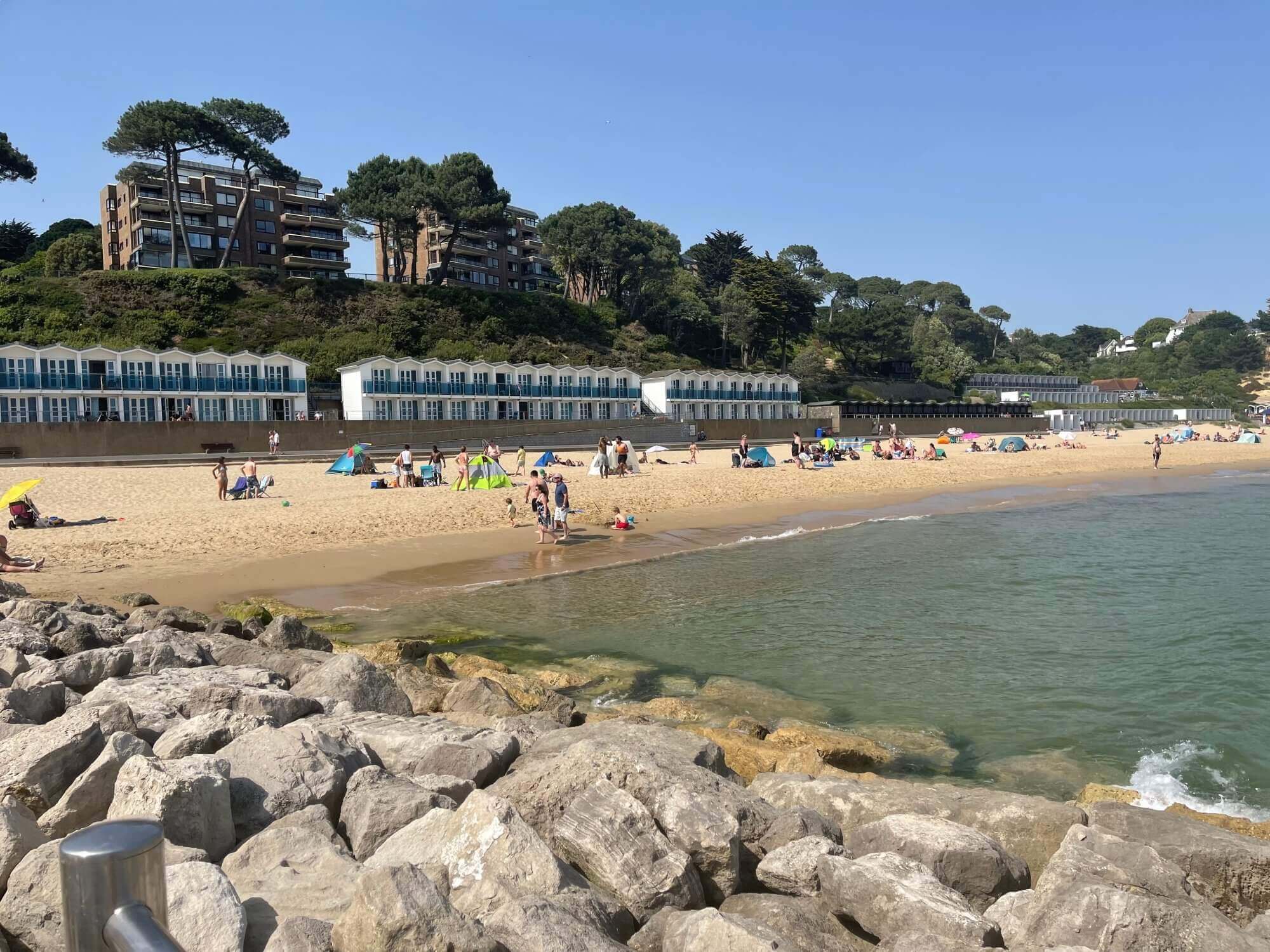 Beachgoers enjoy a sunny day at the beach with people swimming, sunbathing, and beach huts in the background. - Home Instead Poole
