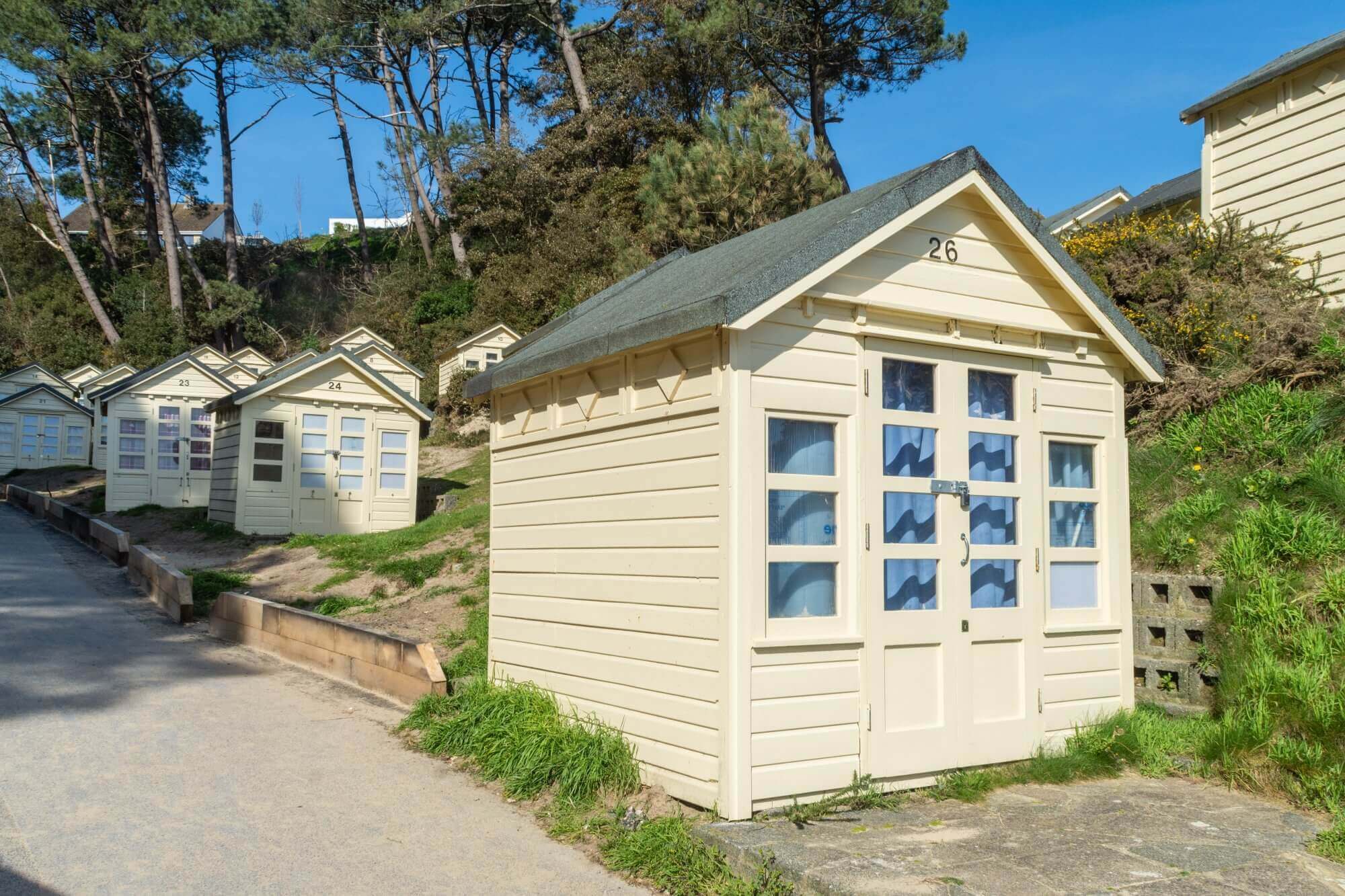 A row of small, cream-coloured beach huts with numbered doors, situated along a path with trees and bushes in the background. - Home Instead Poole