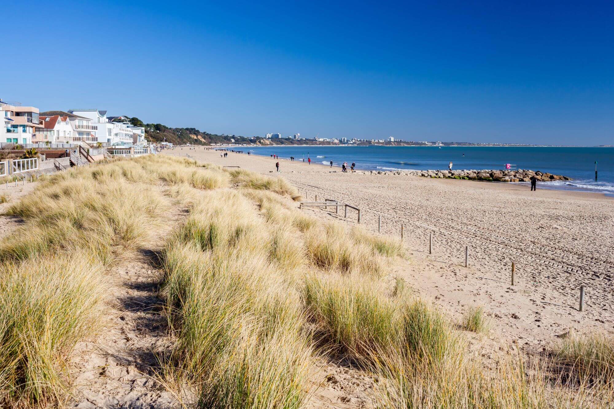 A sunny beach scene with sandy dunes in the foreground, buildings to the left, and people enjoying the beach in the distance. - Home Instead Poole
