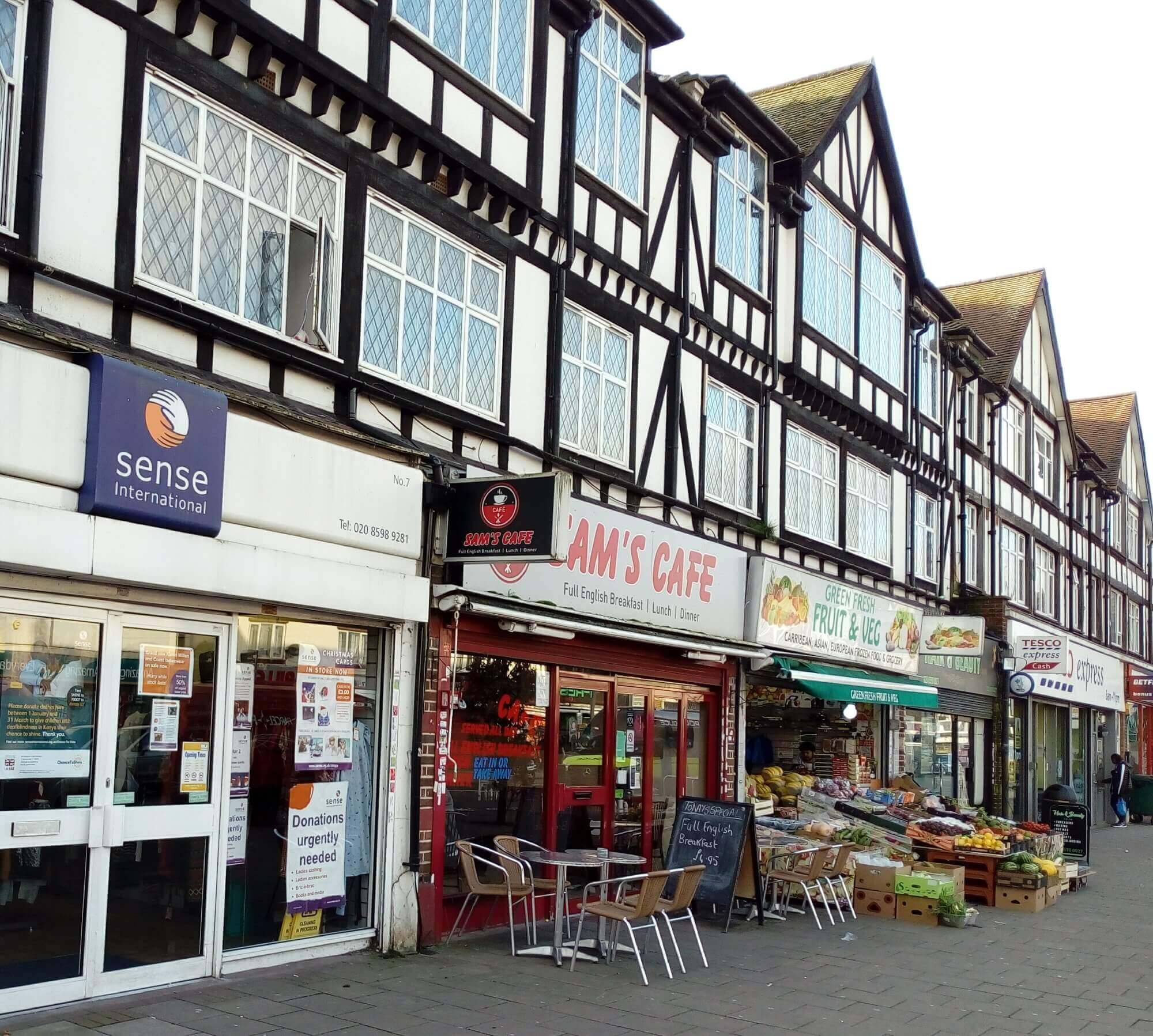 A row of shops, including a charity store, a cafe, a fruit & veg store, and a Tesco Express, on a street. - Home Instead