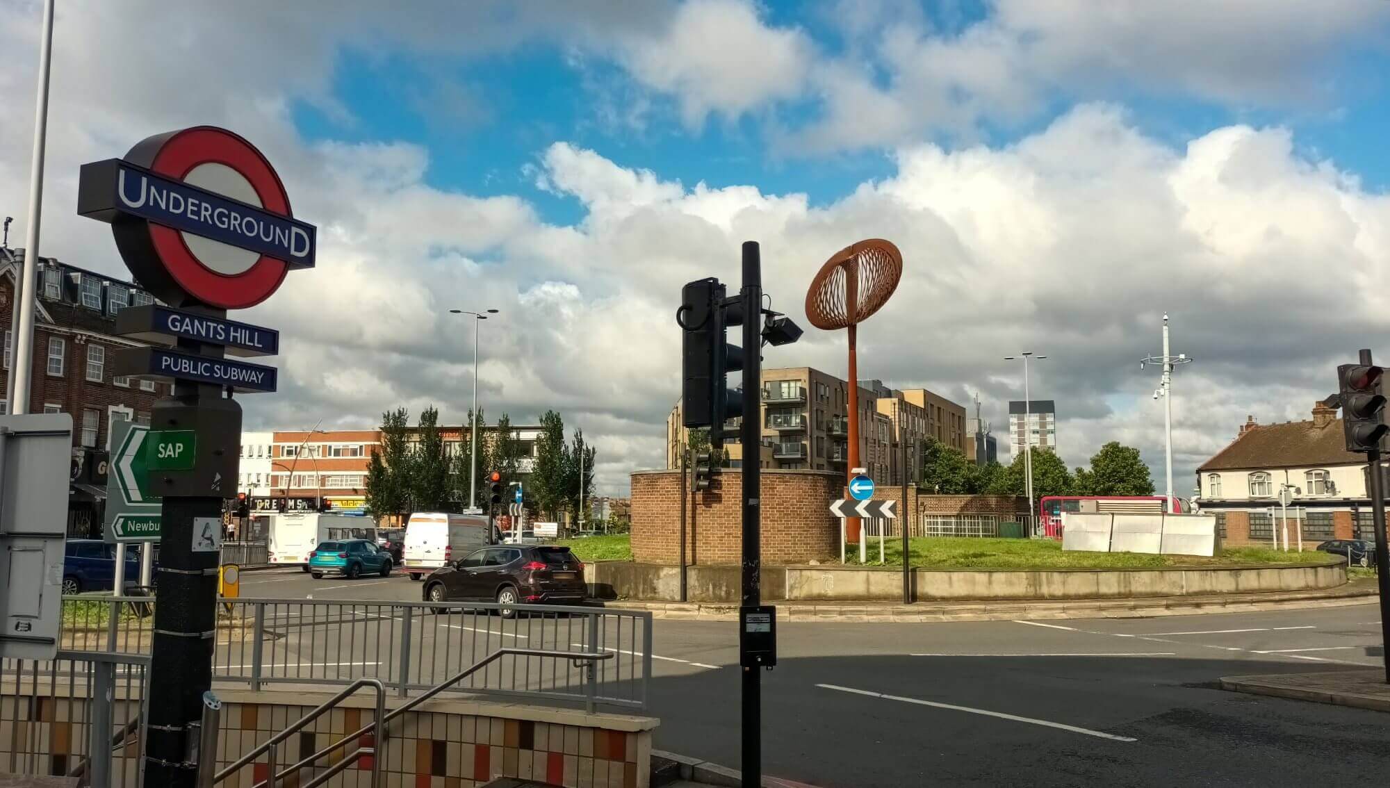 Street scene featuring Gants Hill Underground Station sign, traffic lights, cars, and a unique sculpture on a roundabout. - Home Instead