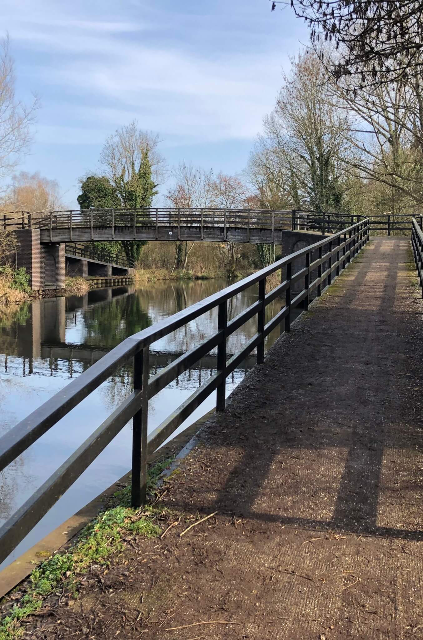 A dirt path with a black railing leads to a wooden footbridge over a calm river, surrounded by trees. - Home Instead