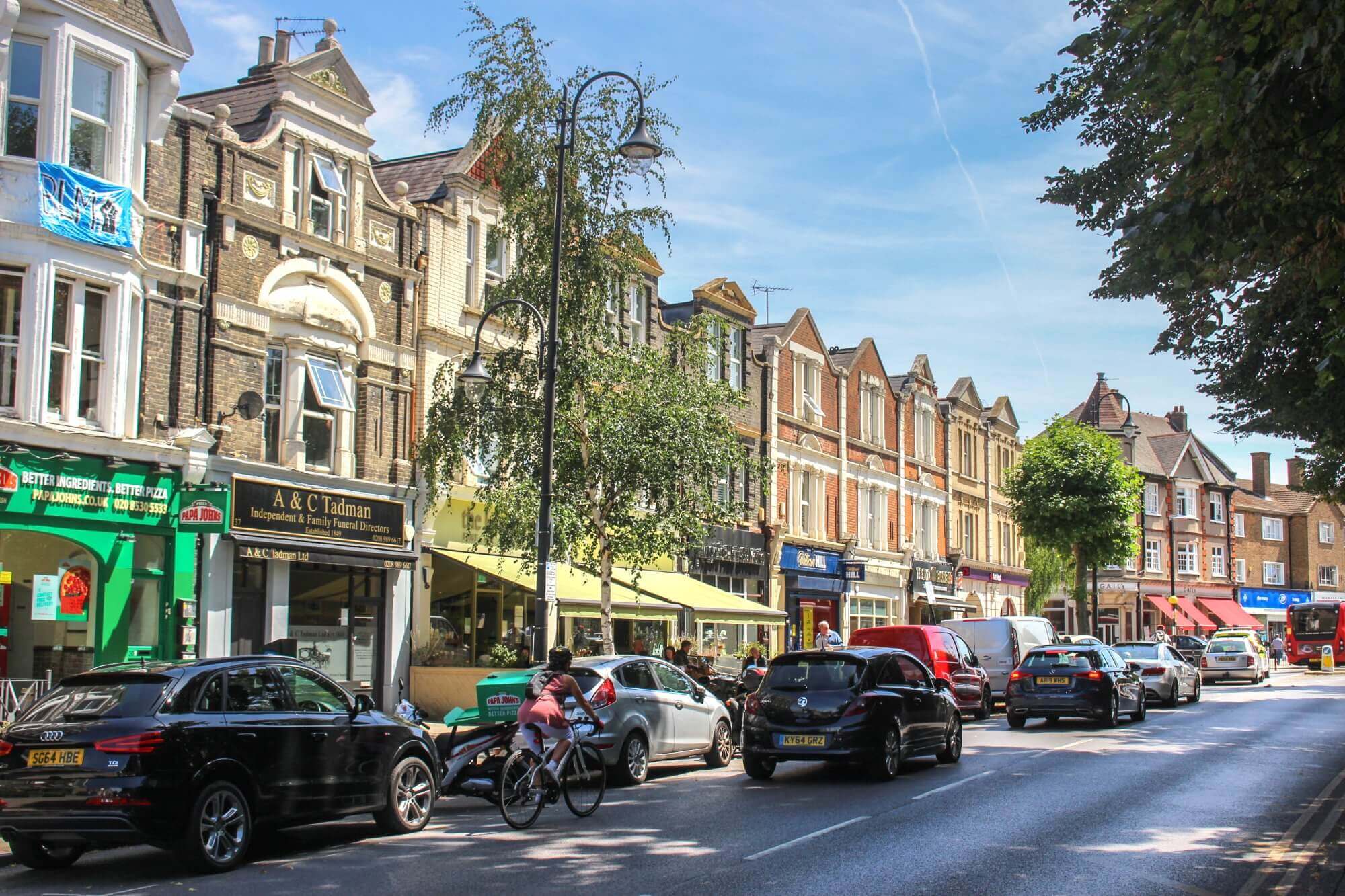 Busy street with parked cars, pedestrians, cyclist, and shops. Victorian-style buildings line the sunny street. - Home Instead