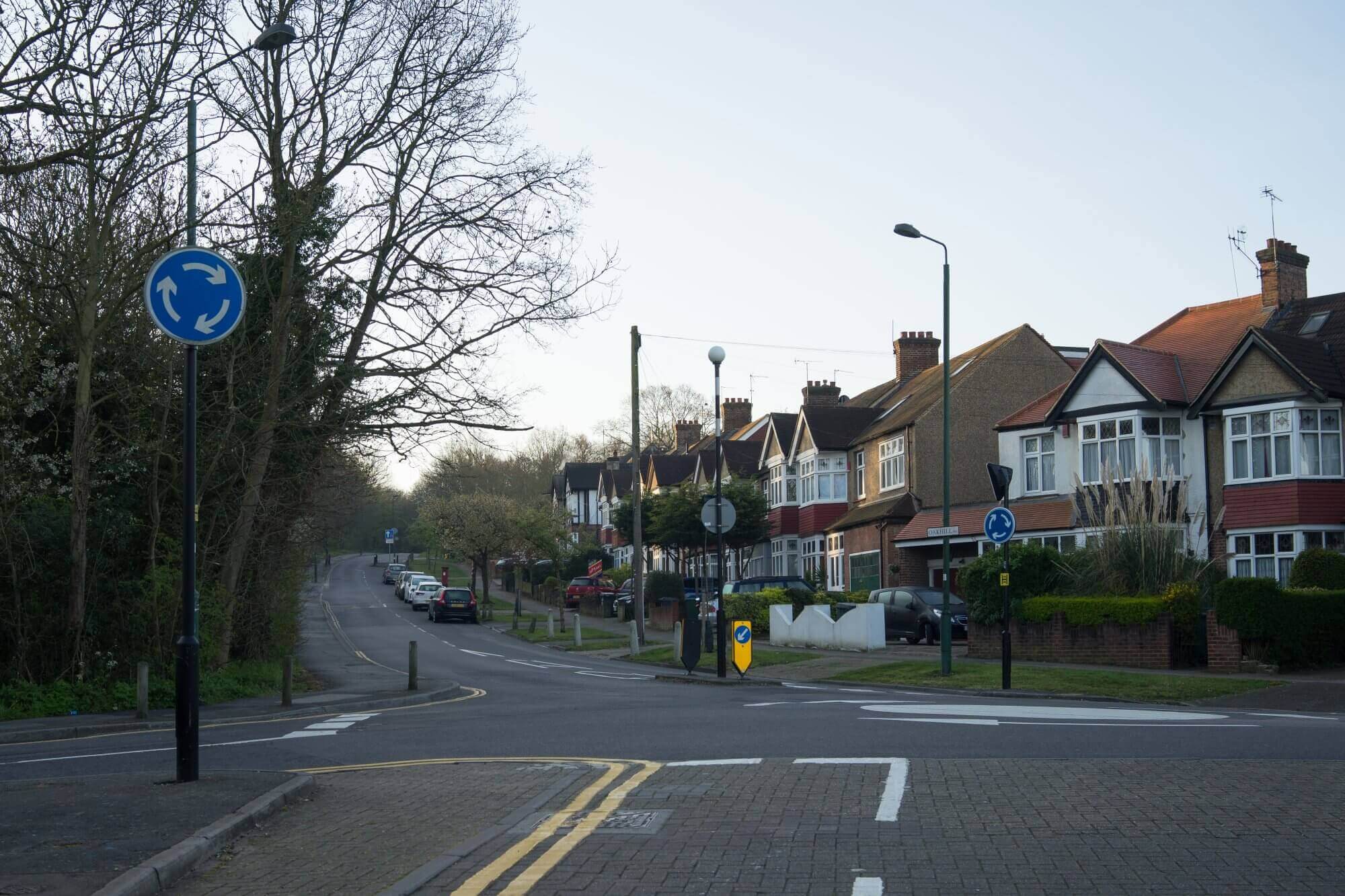 Residential street with houses on the right, a small roundabout with blue signs, and trees on the left side of the road. - Home Instead