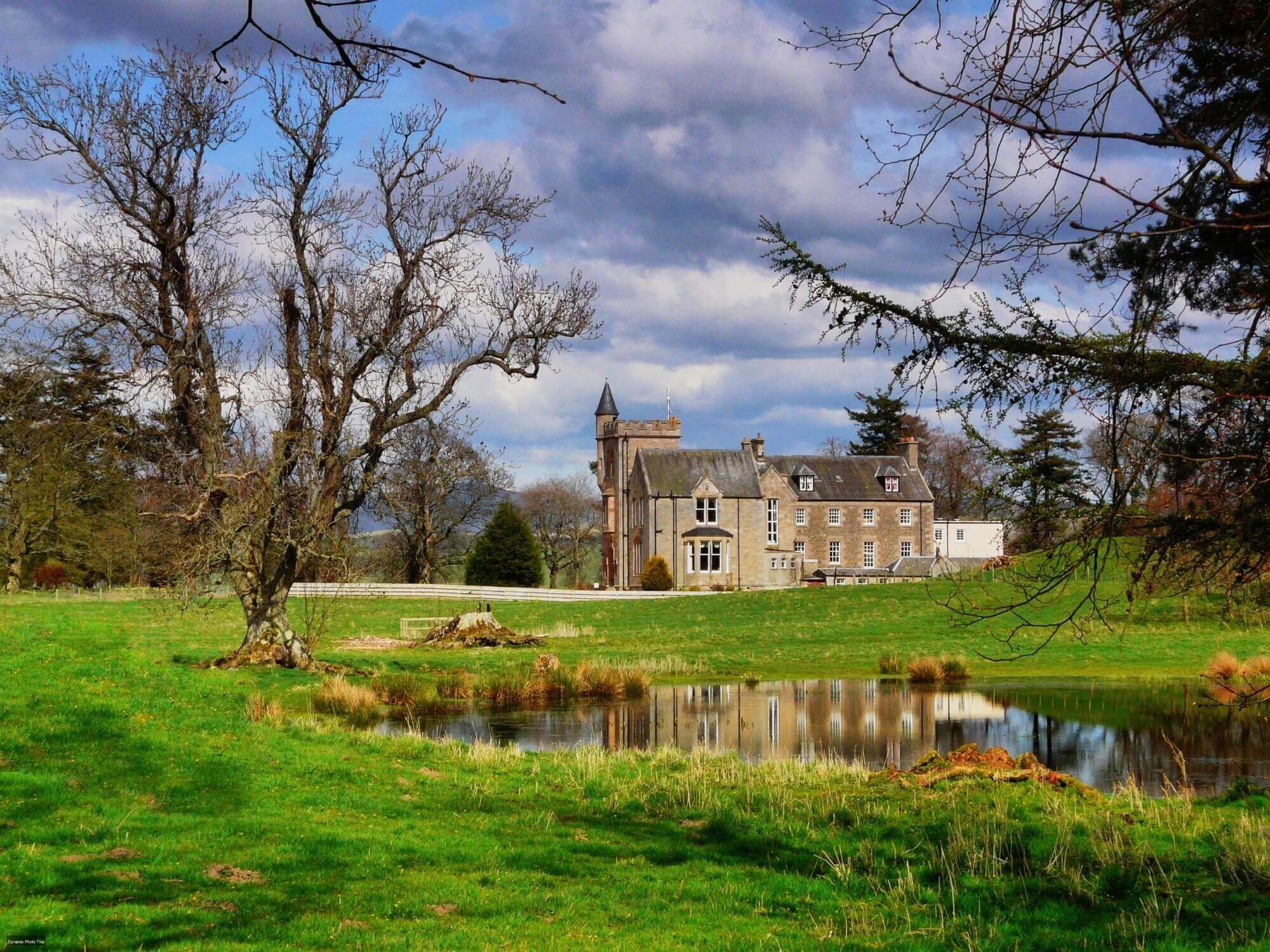 A large, historic stone house with a tower, set on a grassy estate with a pond in the foreground under a partly cloudy sky. - Home Instead