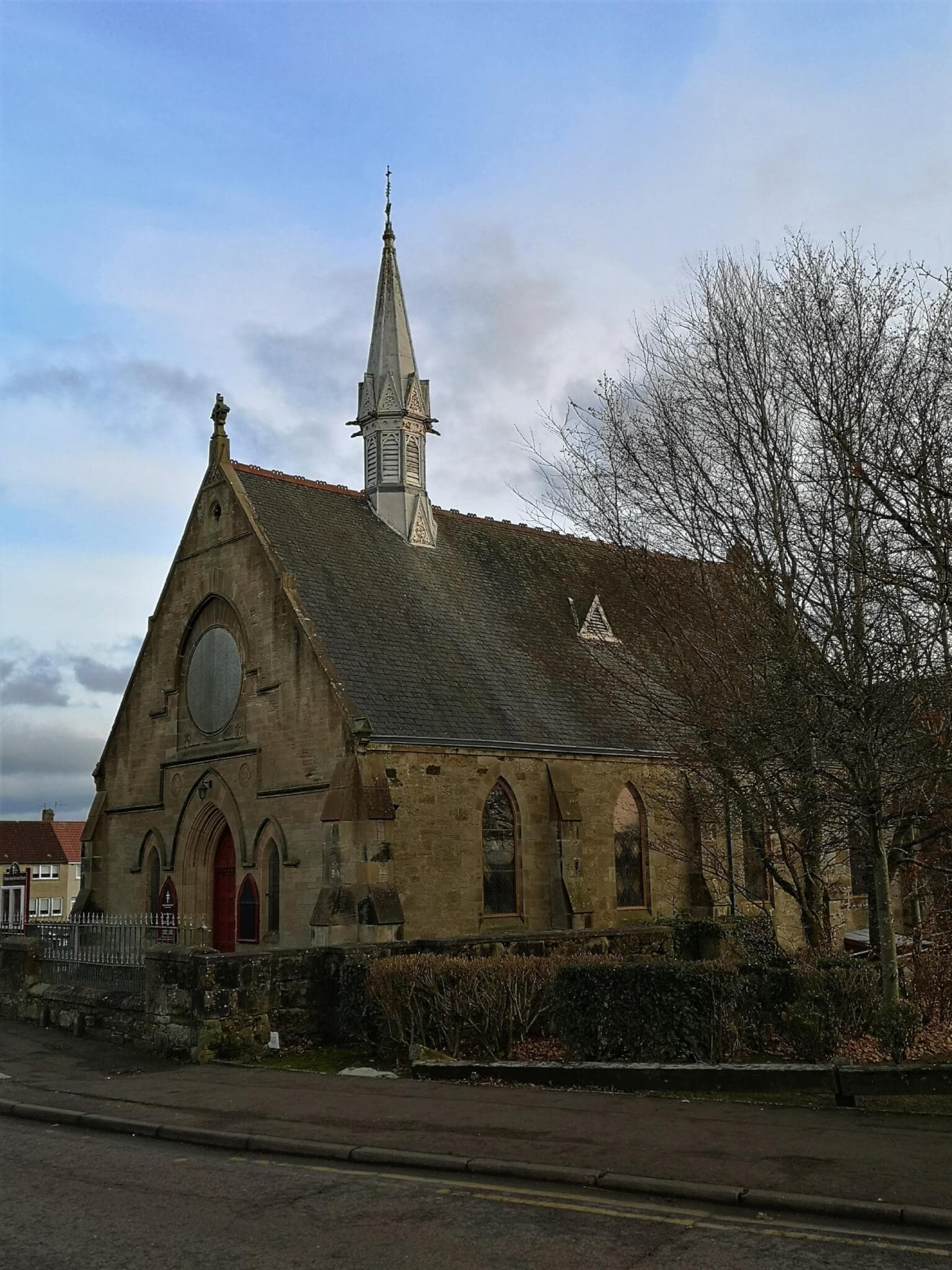 Historic stone church with tall steeple, arched windows, and red doors, set against a cloudy sky. Bare trees in foreground. - Home Instead