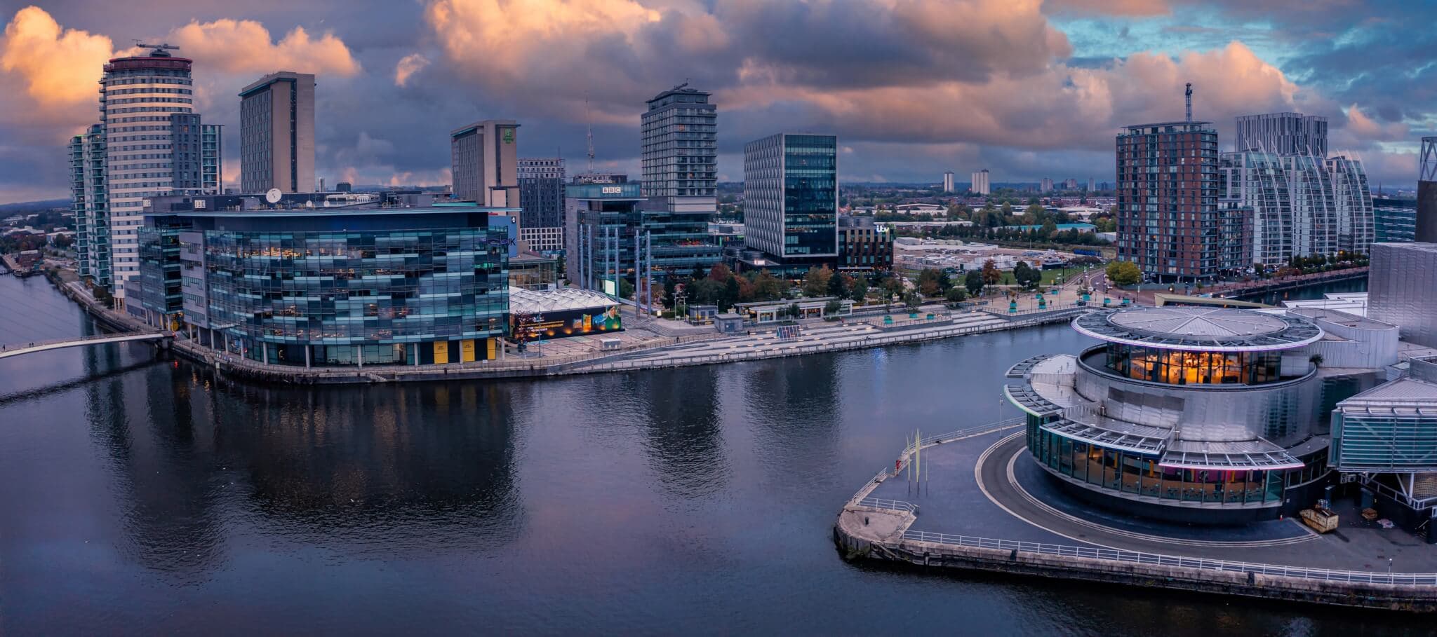 Panoramic view of a modern waterfront city skyline at dusk, with buildings reflecting in calm water under a cloudy sky. - Home Instead