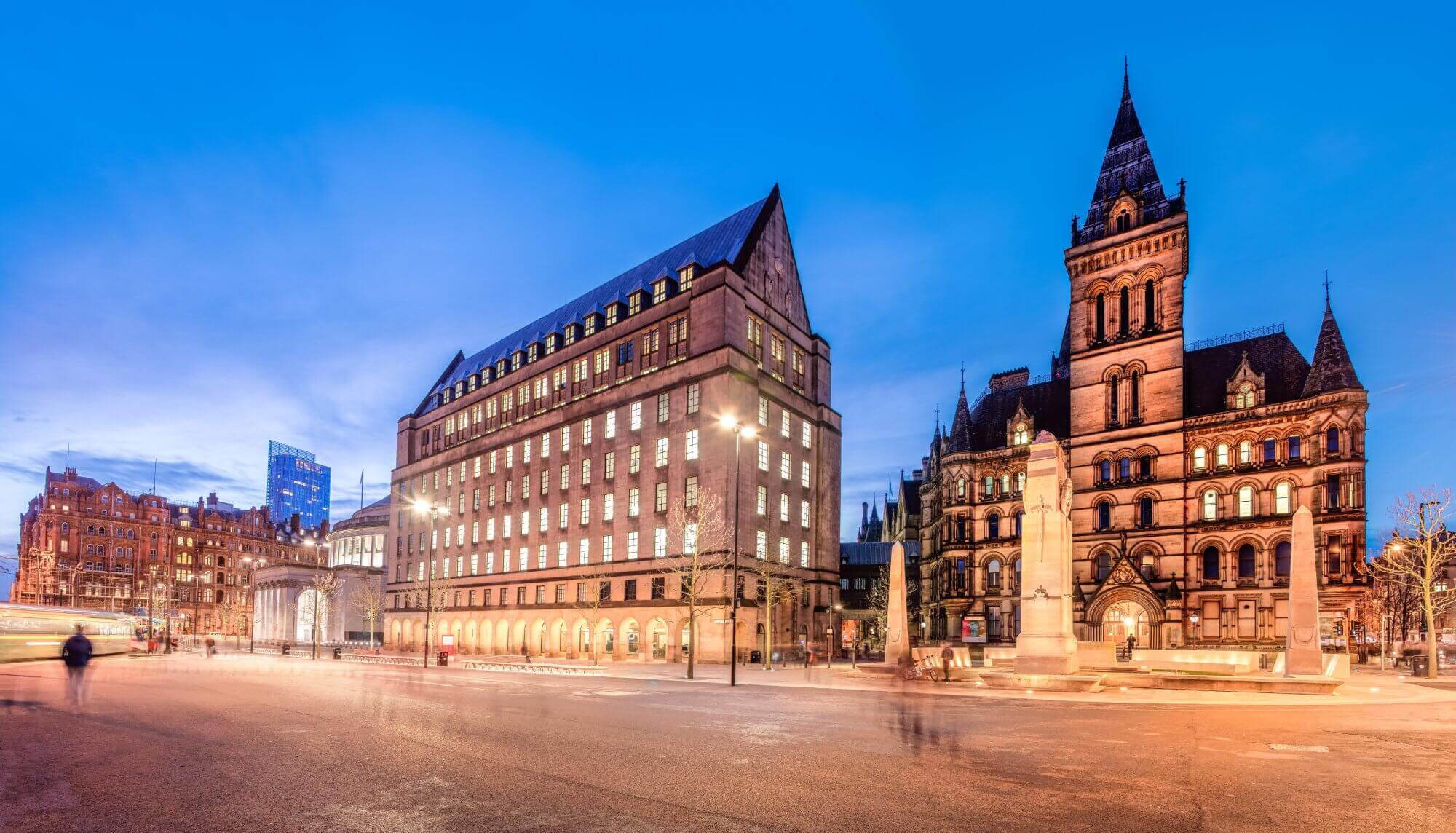 Illuminated historic buildings at dusk in Manchester, with blurred pedestrian motion and a clear blue sky. - Home Instead