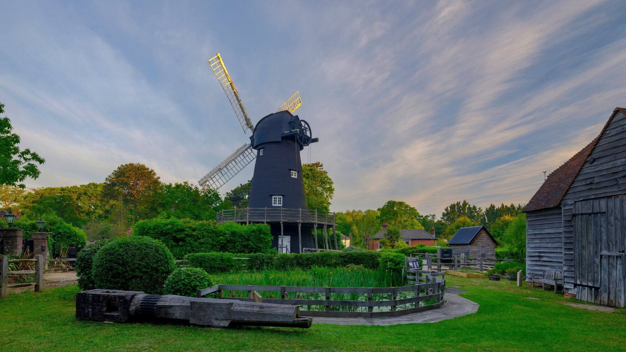 A black windmill under a cloudy sky, surrounded by greenery, wooden buildings, and a wooden trough in the foreground. - Home Instead Southampton