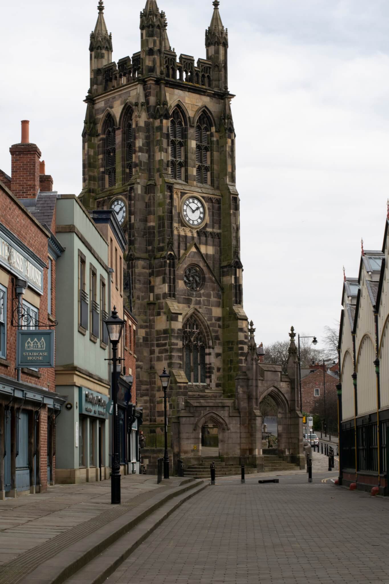 Tall, historic clock tower on a quiet street lined with buildings and shops under a cloudy sky. - Home Instead
